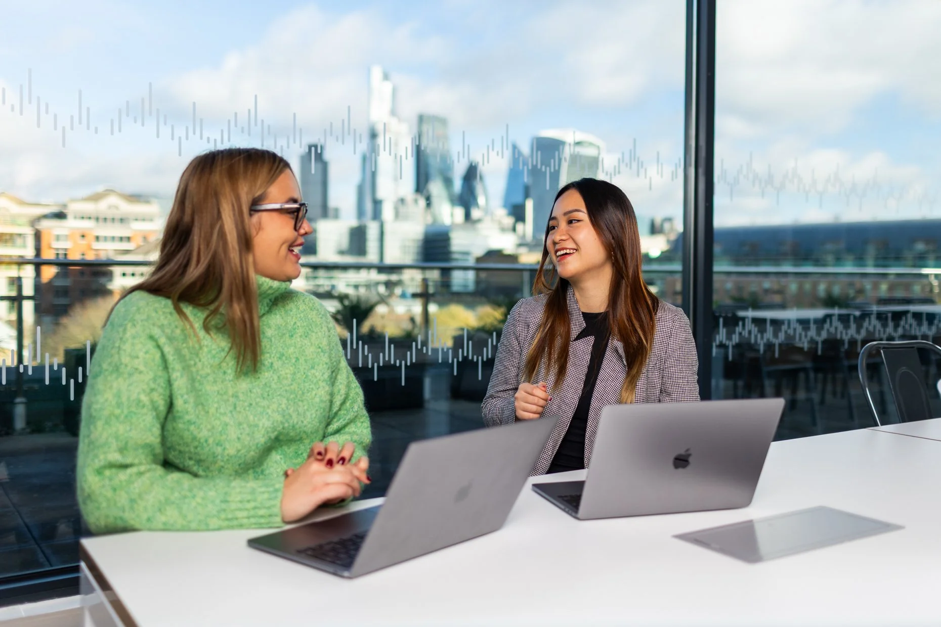 Two women smiling and talking during a meeting in a modern office with a city skyline view behind them, with laptops on the table.