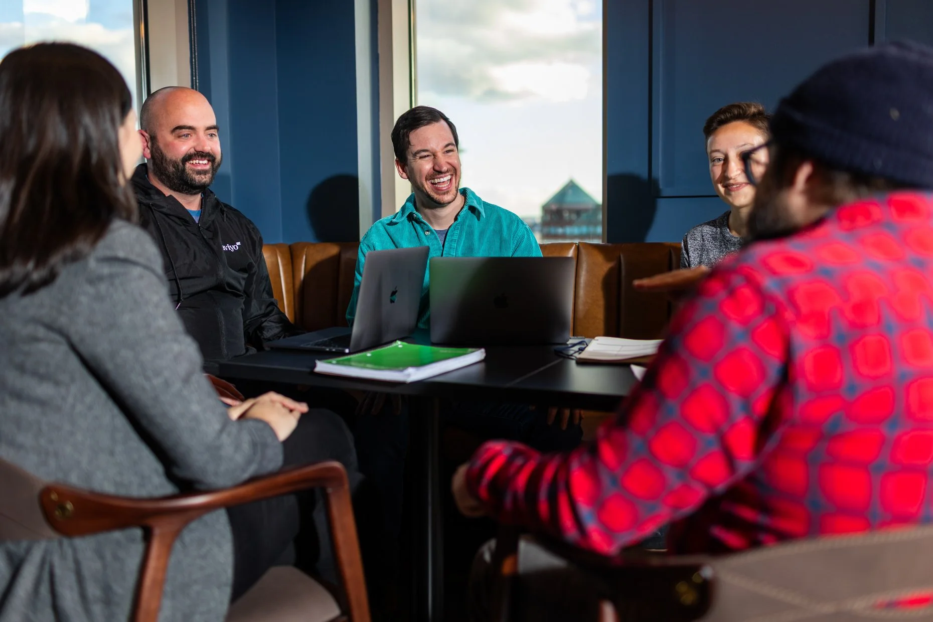 A group of five people sitting around a table in a meeting room, smiling and engaging in conversation. Two laptops, notebooks, and documents are on the table. Large window in the background shows an outdoor scene with a building and cloudy sky.