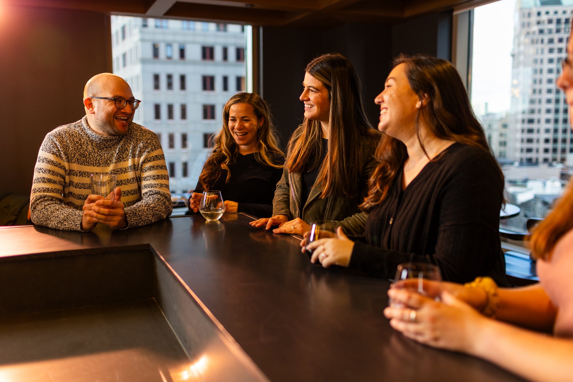 Five people are gathered around a dining table in a city high-rise, enjoying drinks and conversation. They are smiling and appear to be having a good time.