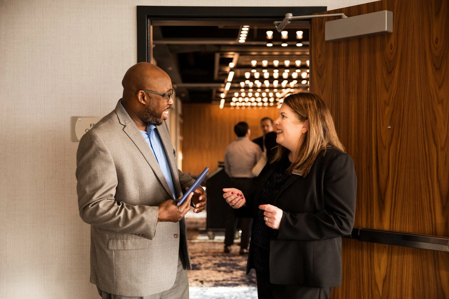 A man and woman in business attire are conversing at a professional event, standing near a wooden wall in an indoor venue.