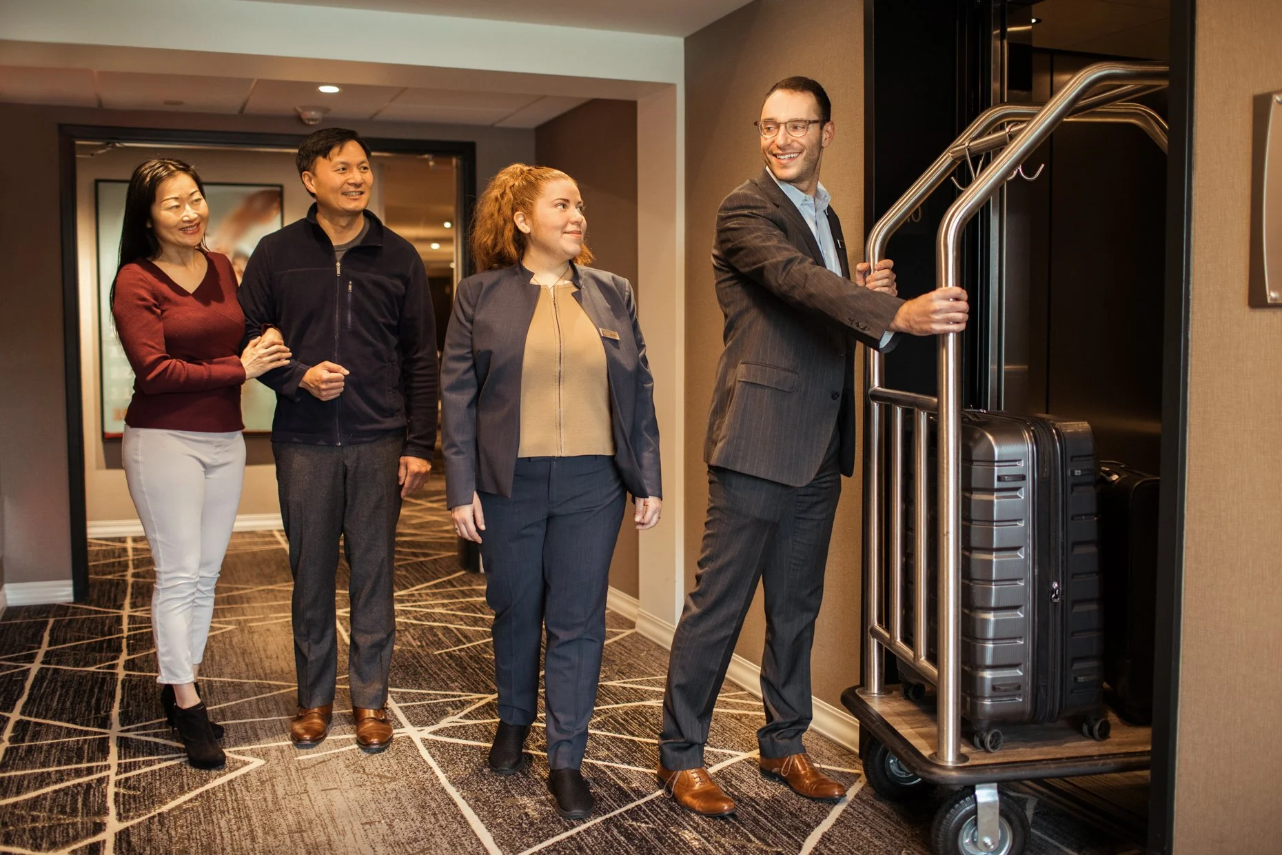 A group of four diverse professionally dressed people standing in line in a hotel hallway, with a hotel bellhop or staff member pushing a luggage cart with suitcases.