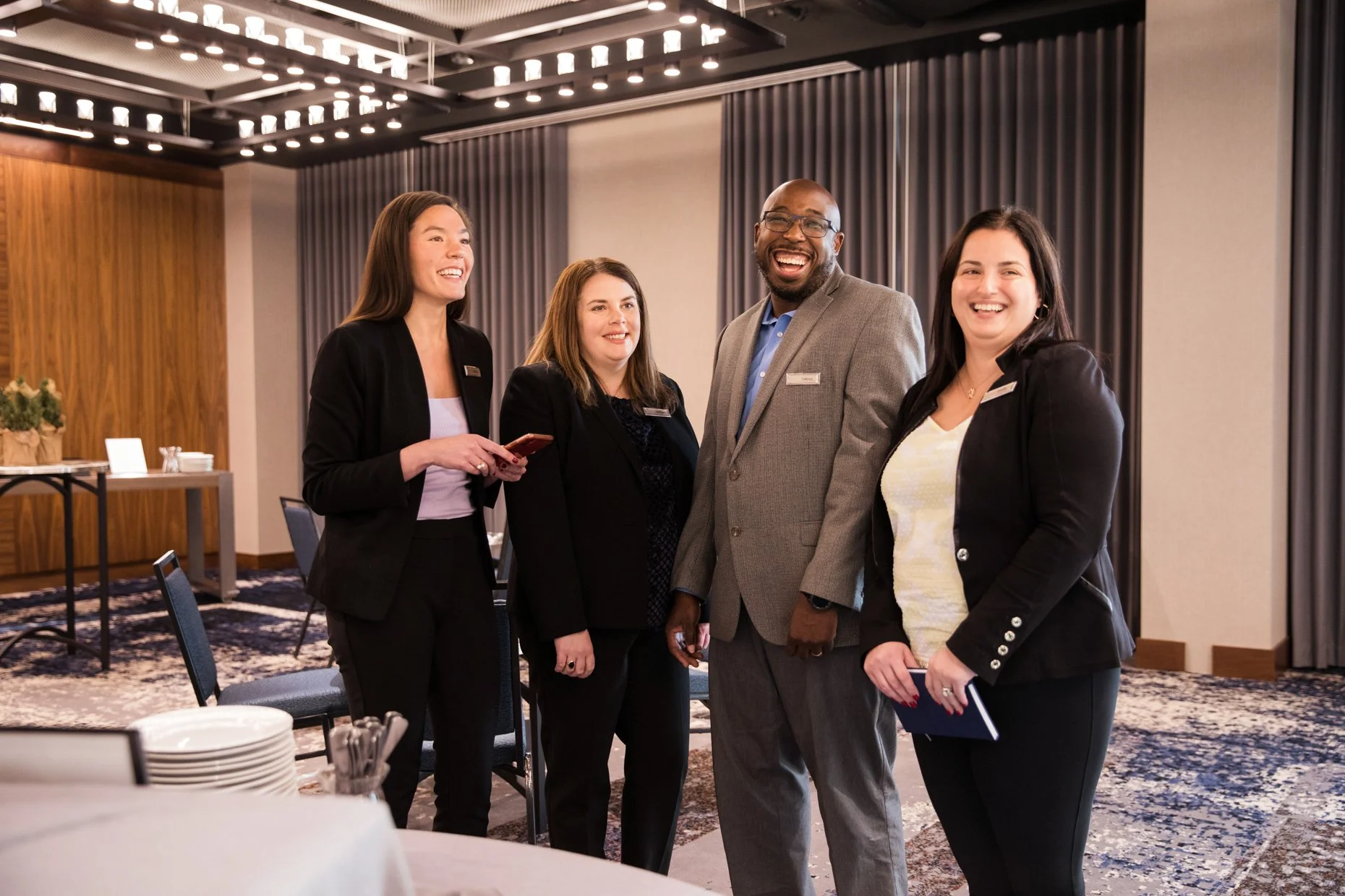 Four diverse professionals standing together and smiling at a formal event in a conference room with round tables, gray curtains, and decorative ceiling lights.