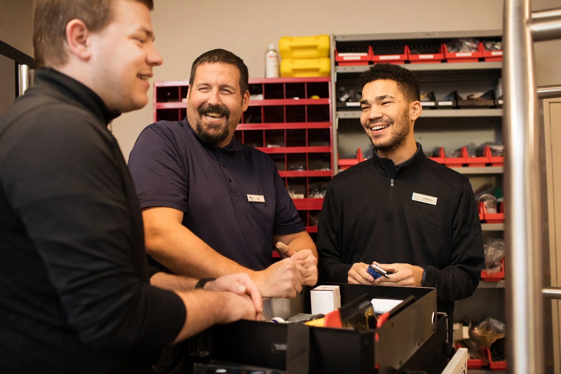 Three men smiling and talking in a workshop or hardware store surrounded by shelves filled with tools and supplies.