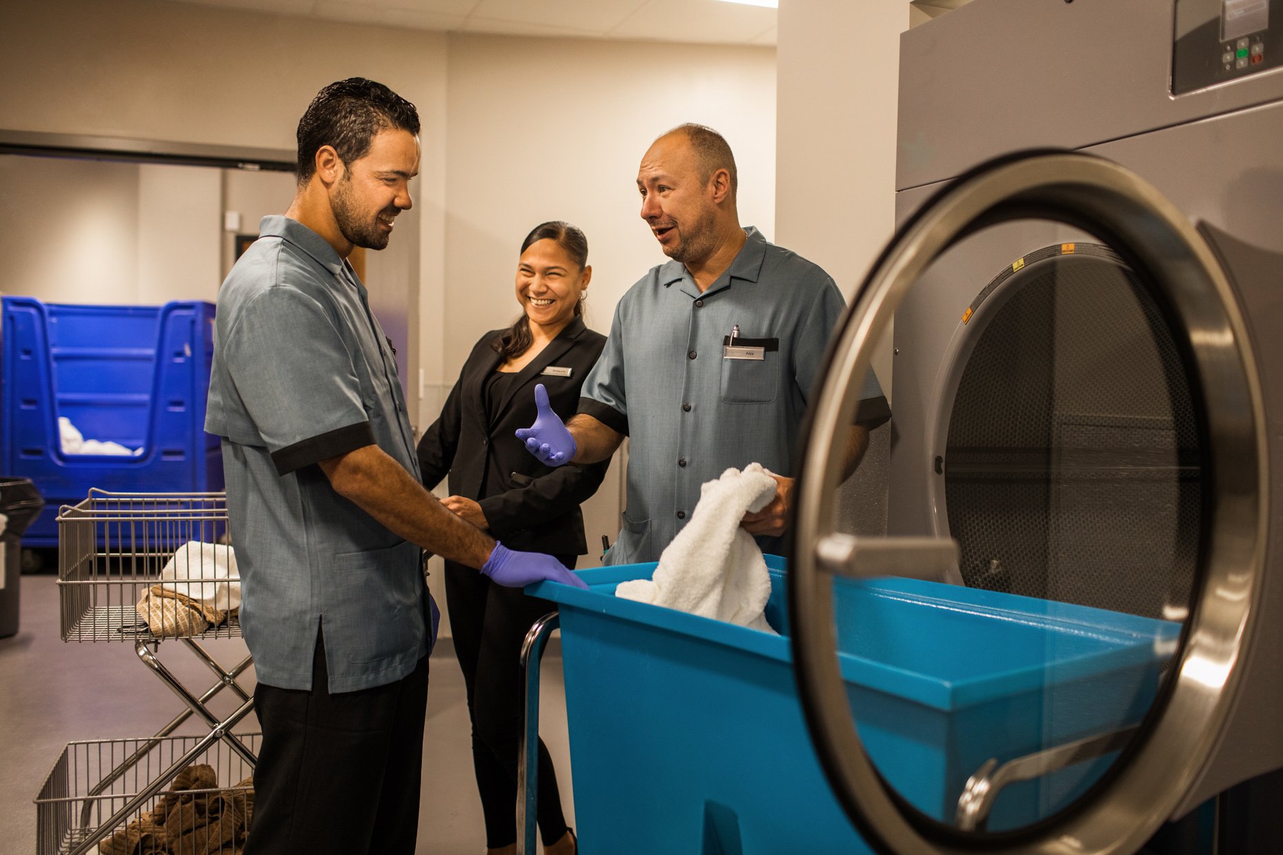 Three hospital staff members in scrubs and a suit smiling and shaking hands in a laundry room with a large industrial washing machine in the foreground.