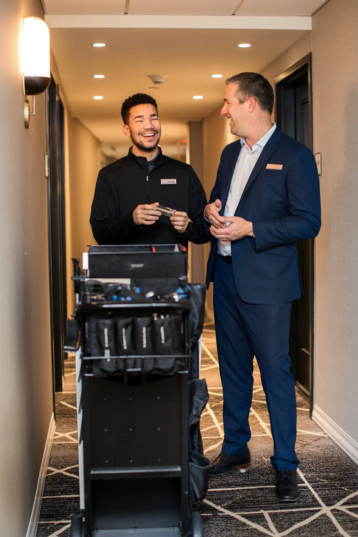 Two men, one in a black jacket and the other in a dark suit, are smiling and talking in a hotel hallway, with a luggage cart in front of them.