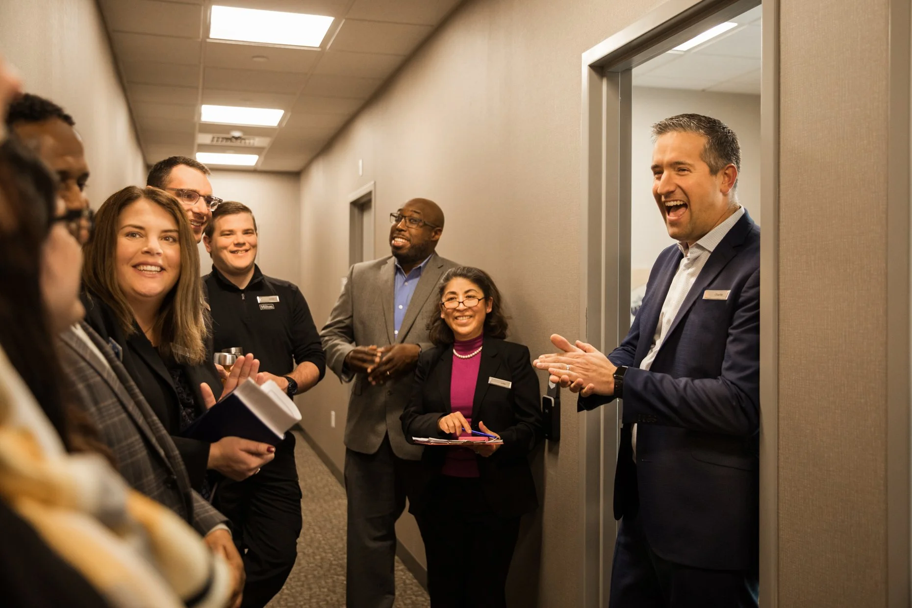 A diverse group of people, including men and women of different ages and ethnicities, are gathered in a hallway, engaging in conversation. One man is smiling and clapping as he speaks. The others are listening, some holding notebooks or papers, and a