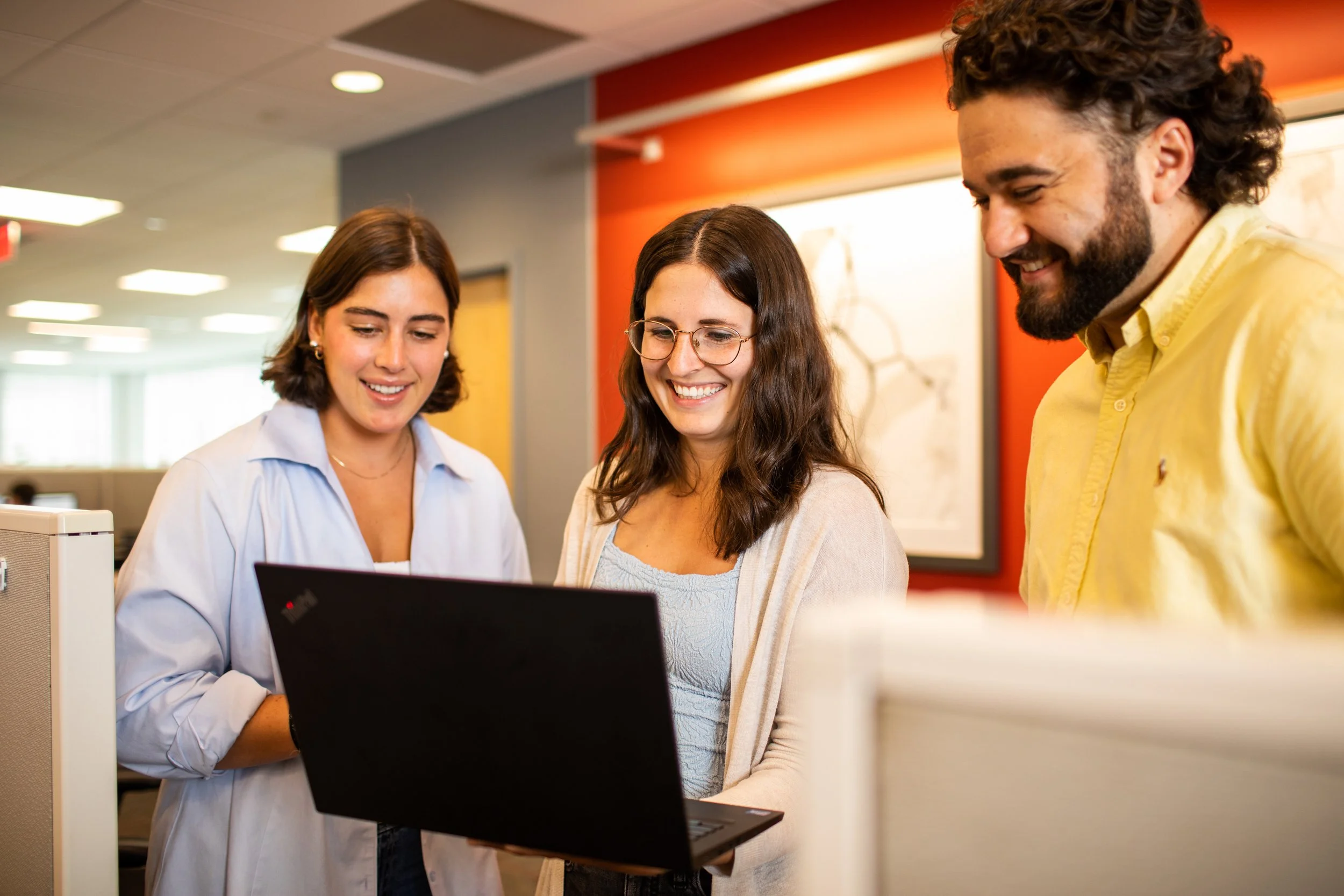 Three people smiling and looking at a laptop screen in an office setting.