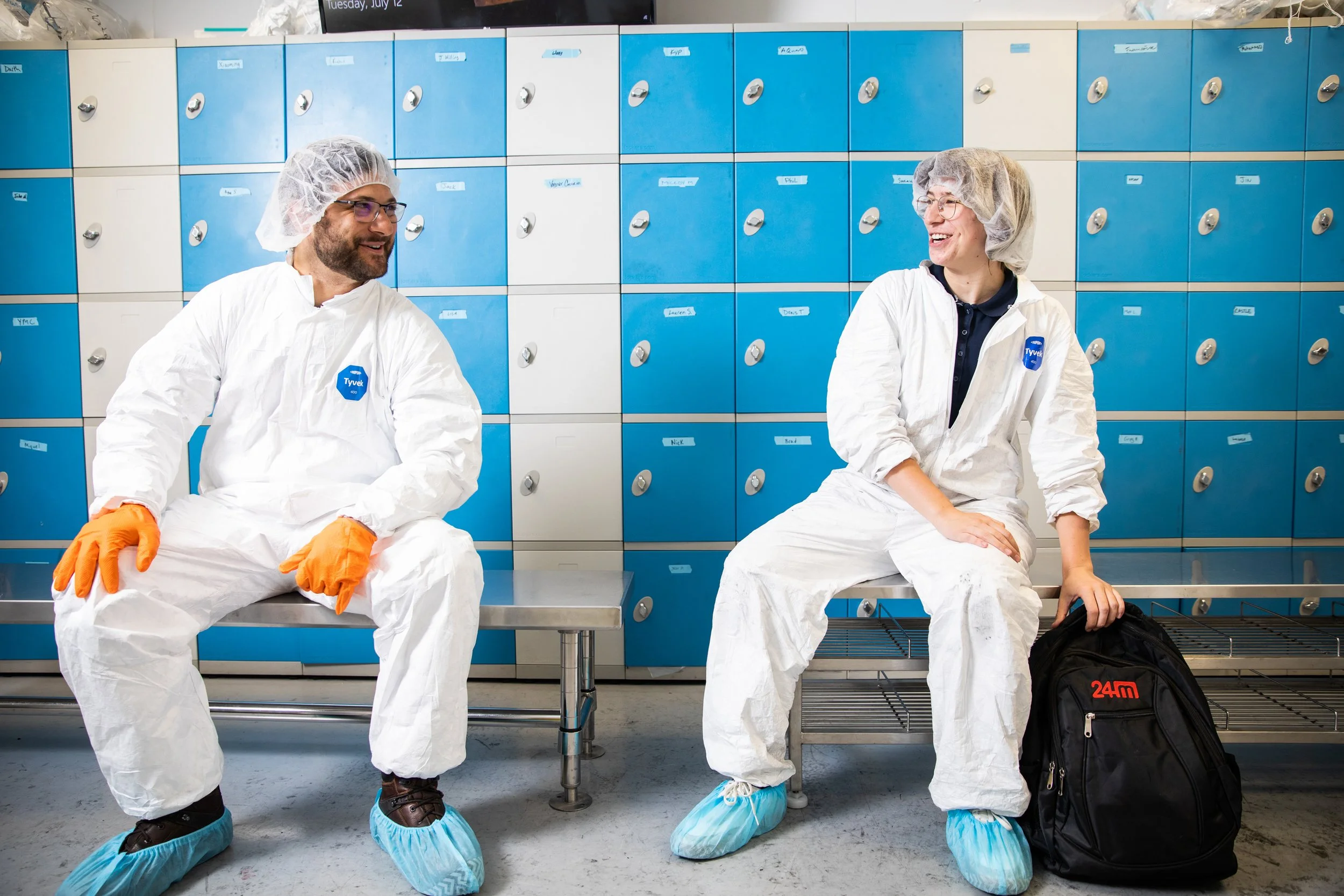Two people wearing white protective suits, hairnets, shoe covers, and gloves, sitting on a bench in front of blue lockers in a locker room. They are smiling and talking to each other.