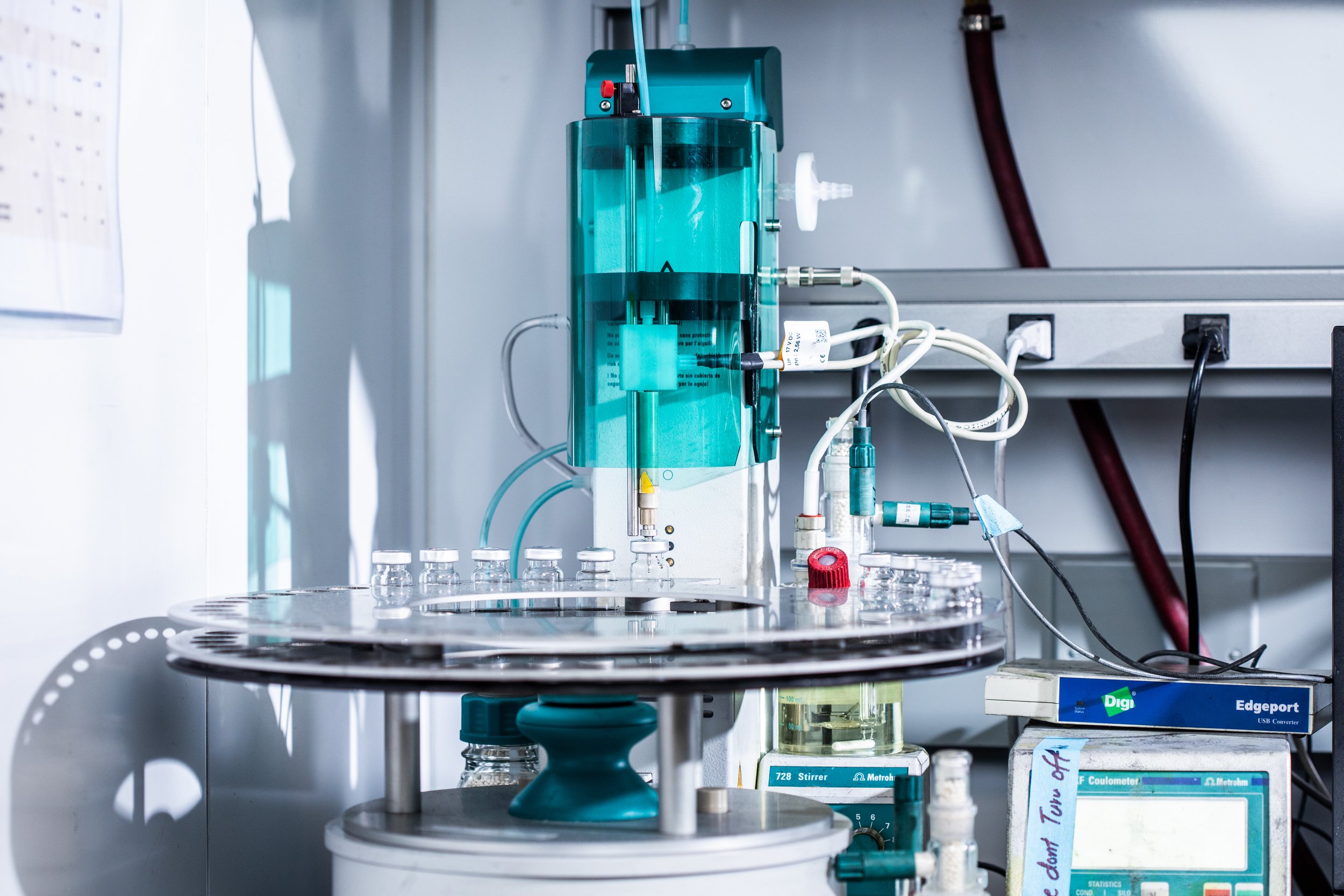 Laboratory equipment including glass bottles on a rotating tray, connected to a turquoise-colored machine with numerous tubes and wires, set on a metal shelf with various digital devices and labels.