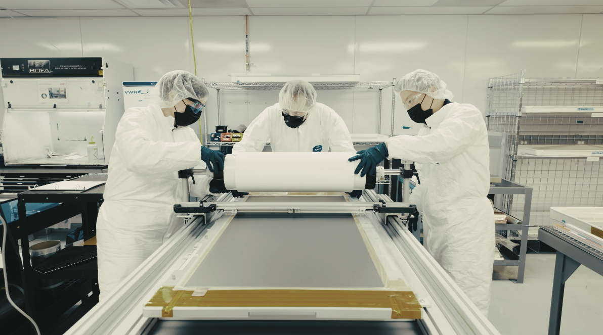 Scientists in lab suits, masks, and gloves handling materials in a cleanroom laboratory environment, working with large sheets of material on a specialized workstation.