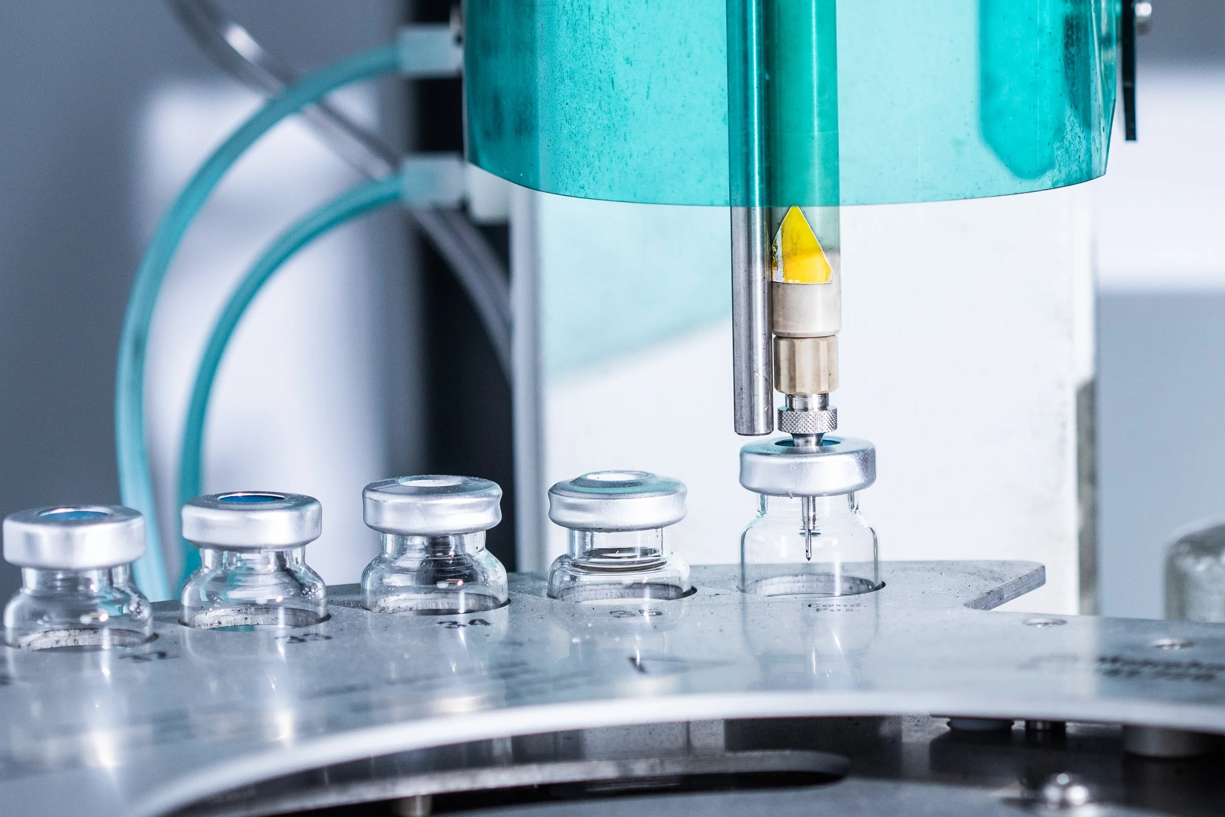 Laboratory machine filling glass vials with liquid, part of pharmaceutical manufacturing process.