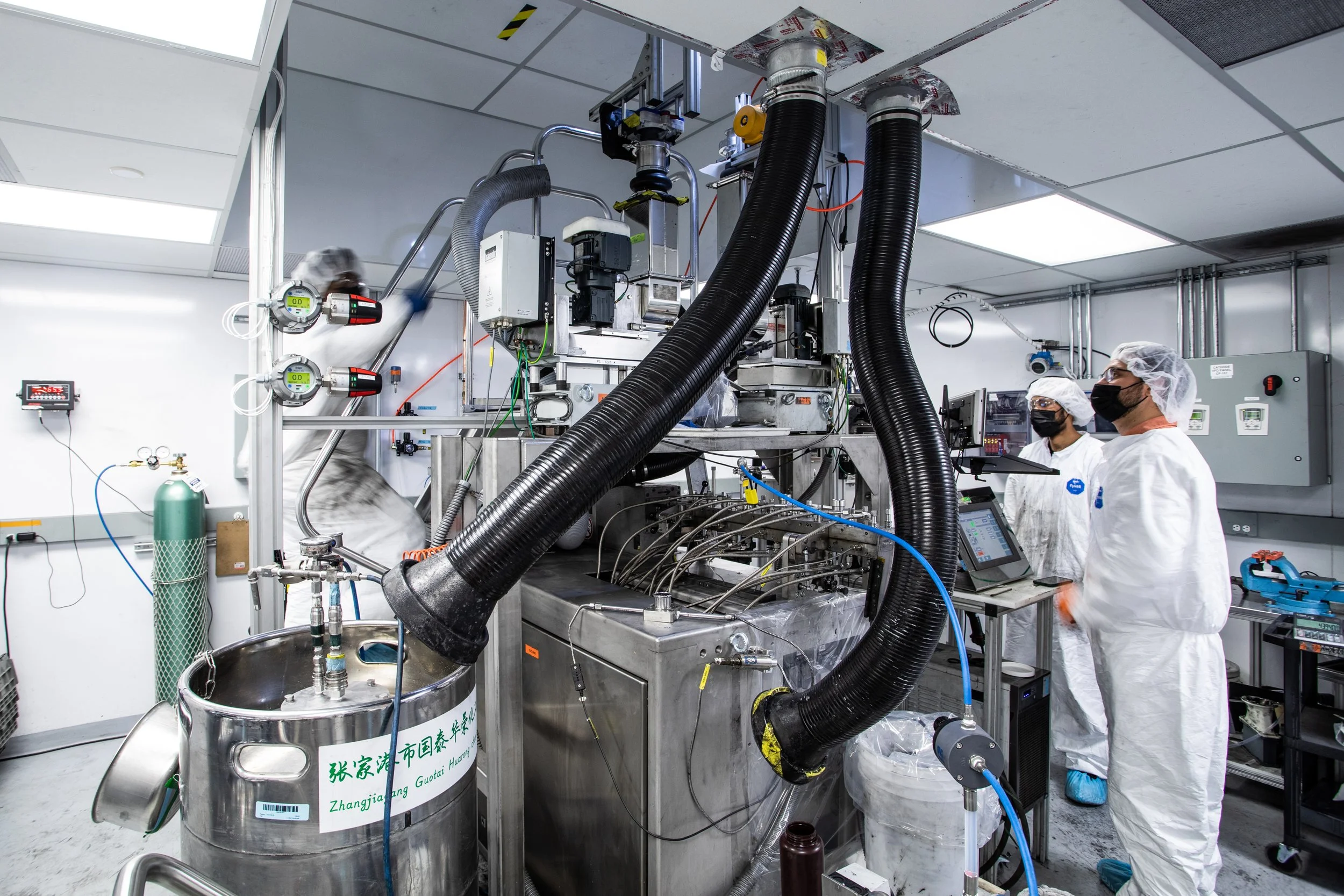 Scientists or engineers in protective clothing and masks working in a high-tech laboratory with machinery, pipes, and digital equipment.