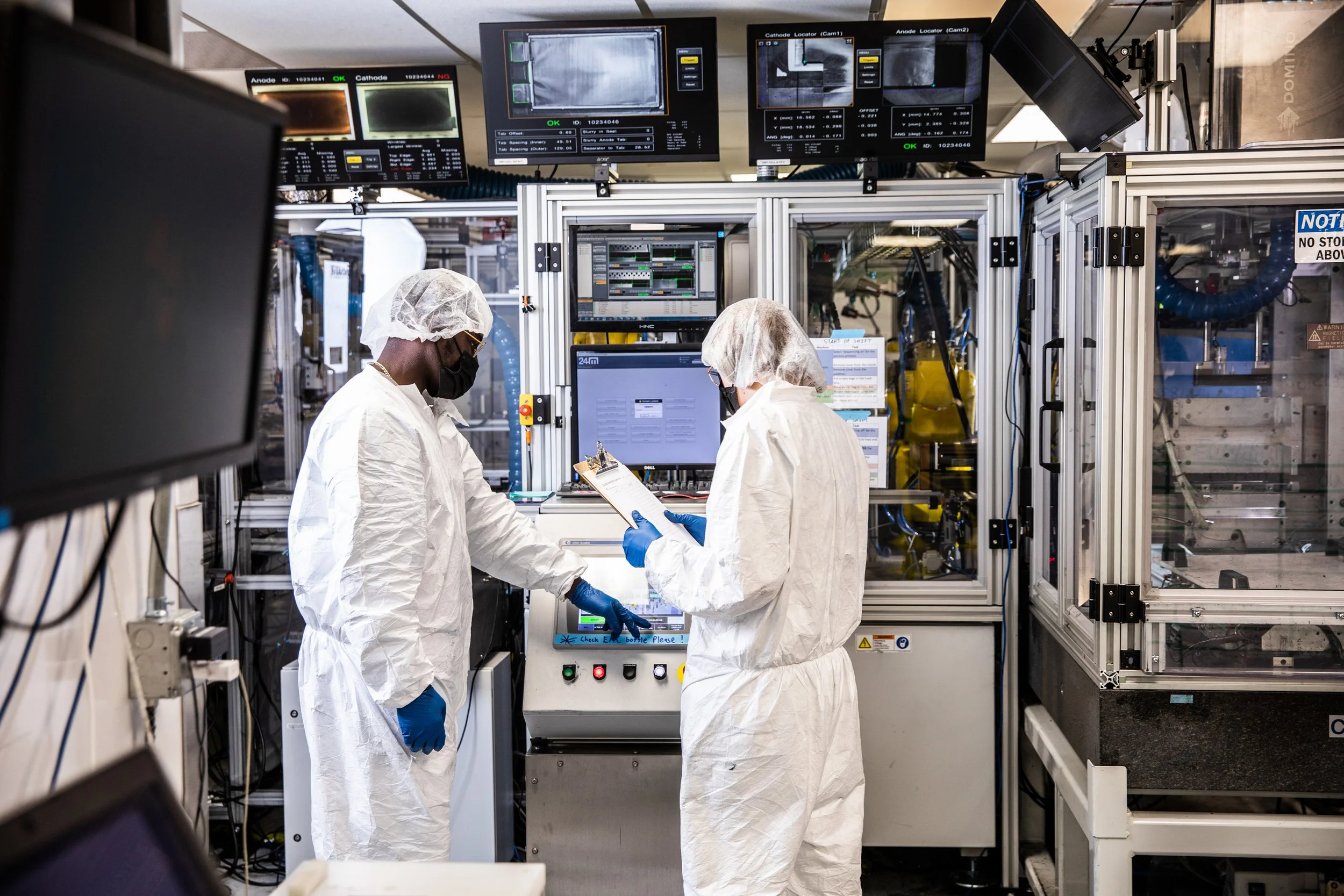 Two scientists wearing white lab coats, hair covers, gloves, and face masks working in a laboratory with electronic equipment and monitors displaying data and images.