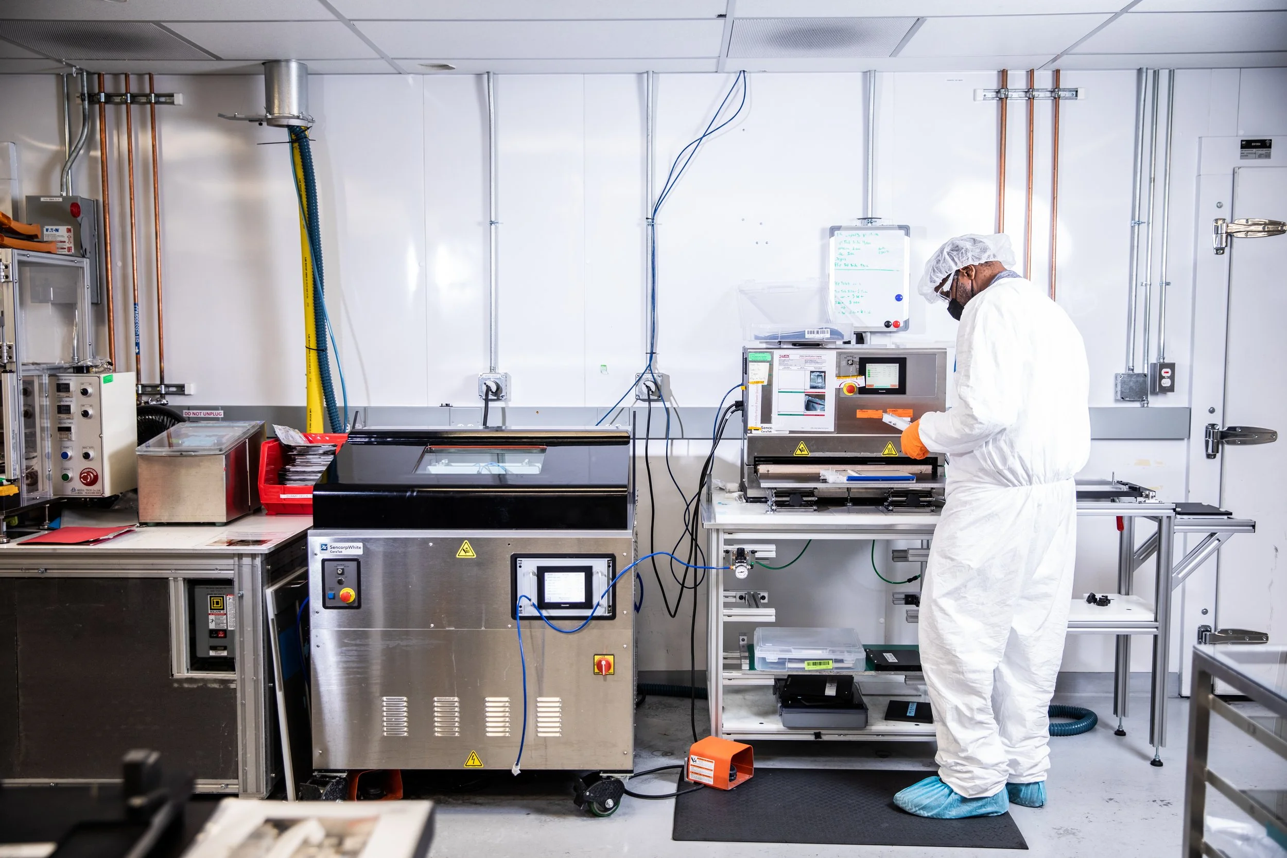 A scientist wearing a white protective suit, hairnet, face mask, gloves, and shoe covers works in a laboratory with various machines and equipment, including clear containers and electronic devices.