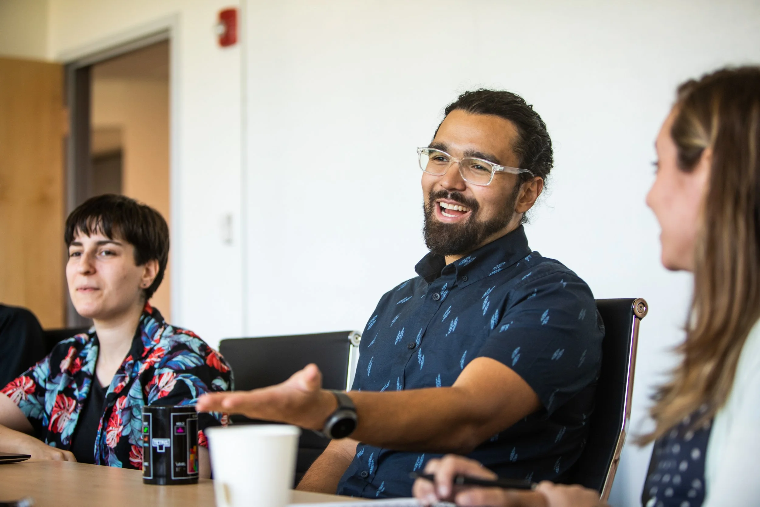 A group of people sitting around a table in a meeting or discussion, with one man wearing glasses and a dark patterned shirt smiling and talking.
