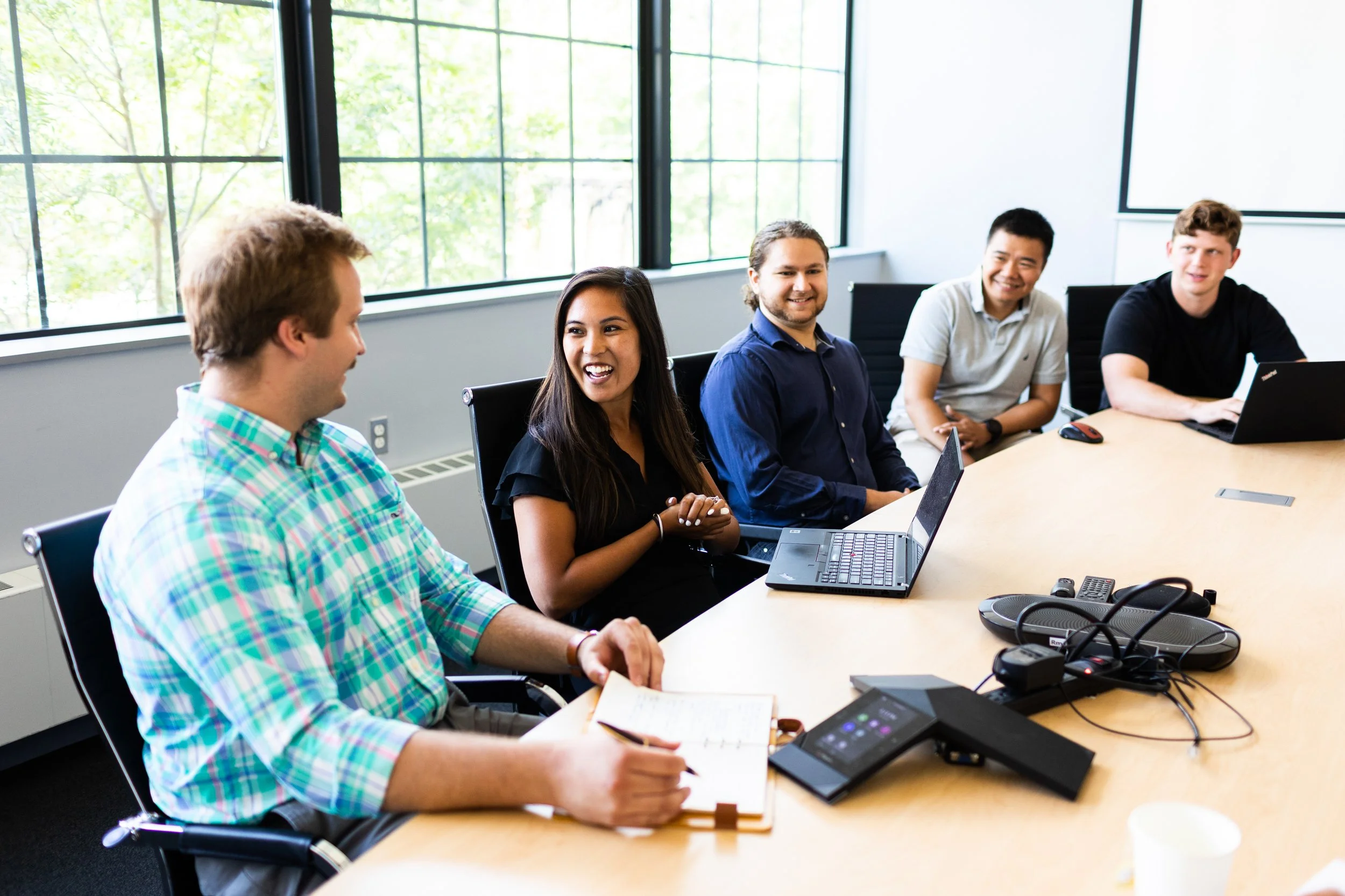 A diverse group of five young adults sitting around a conference table in a modern office, engaging in a discussion or meeting. There are laptops, notebooks, and conference phone equipment on the table. Large windows in the background let in natural 