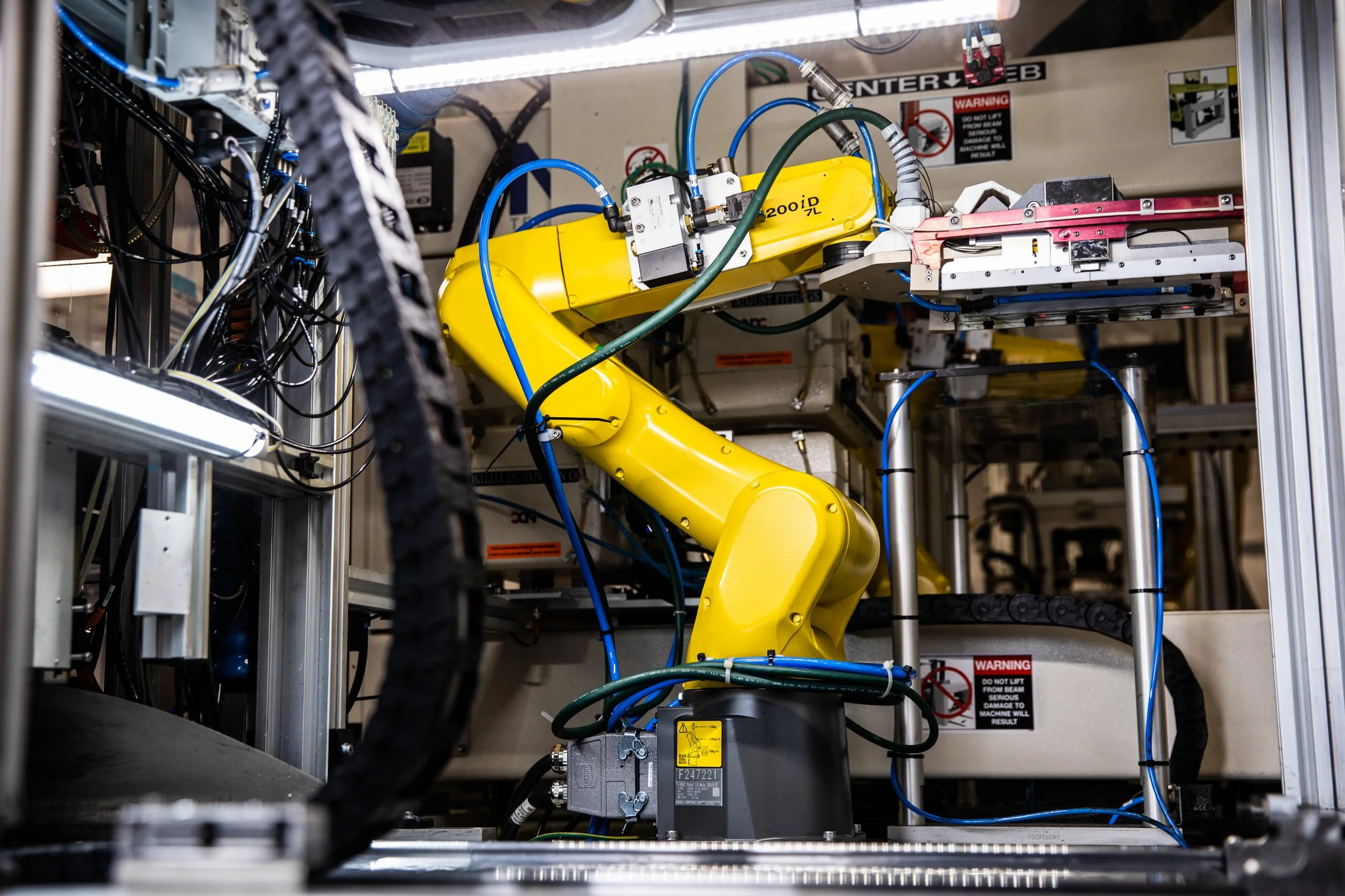 Close-up of industrial robotic arm with yellow cover, blue and green tubes, and various electrical components within a manufacturing or research setting.