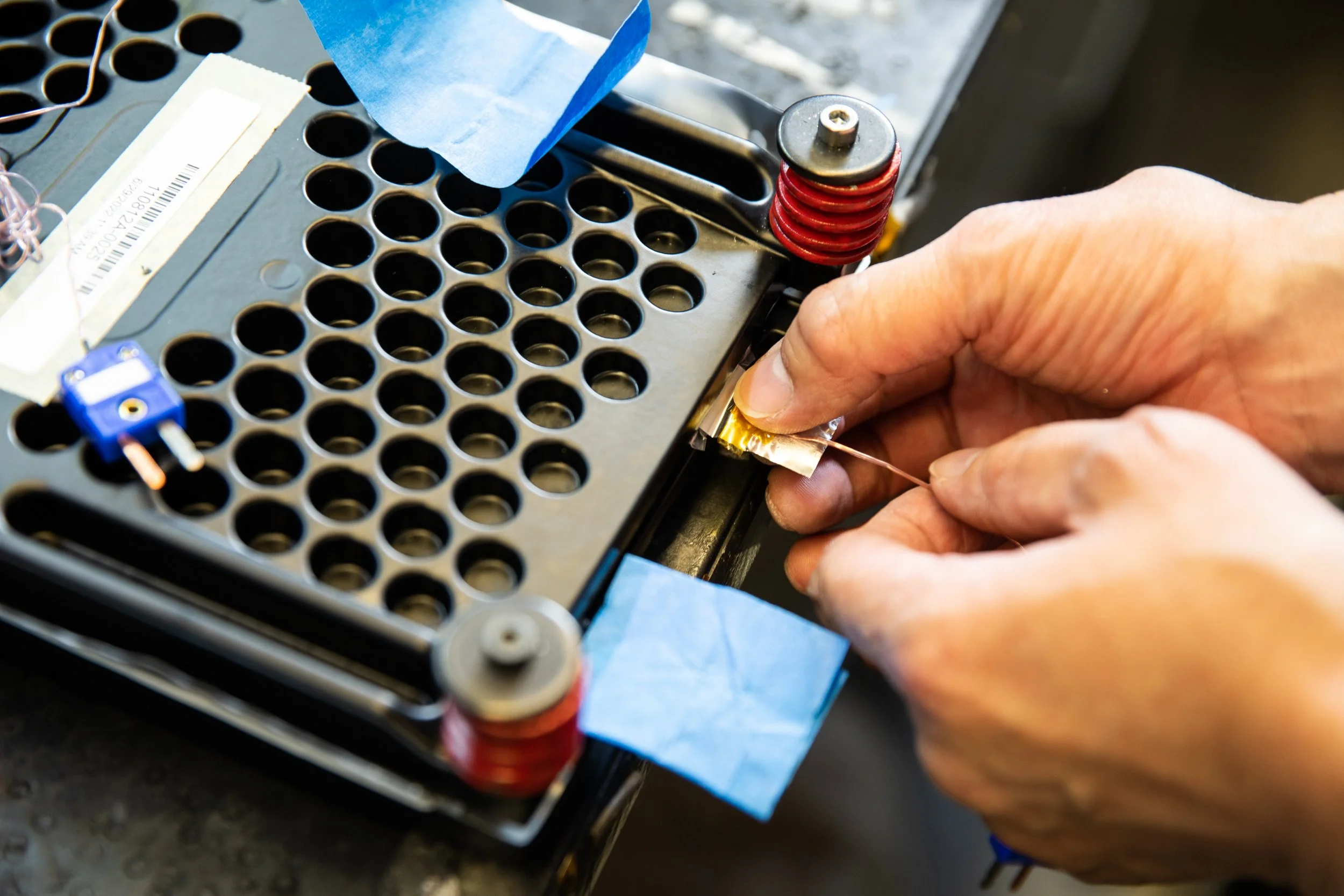 Person's hands working on an electronic device, possibly a circuit board or module, with various components and tools visible.