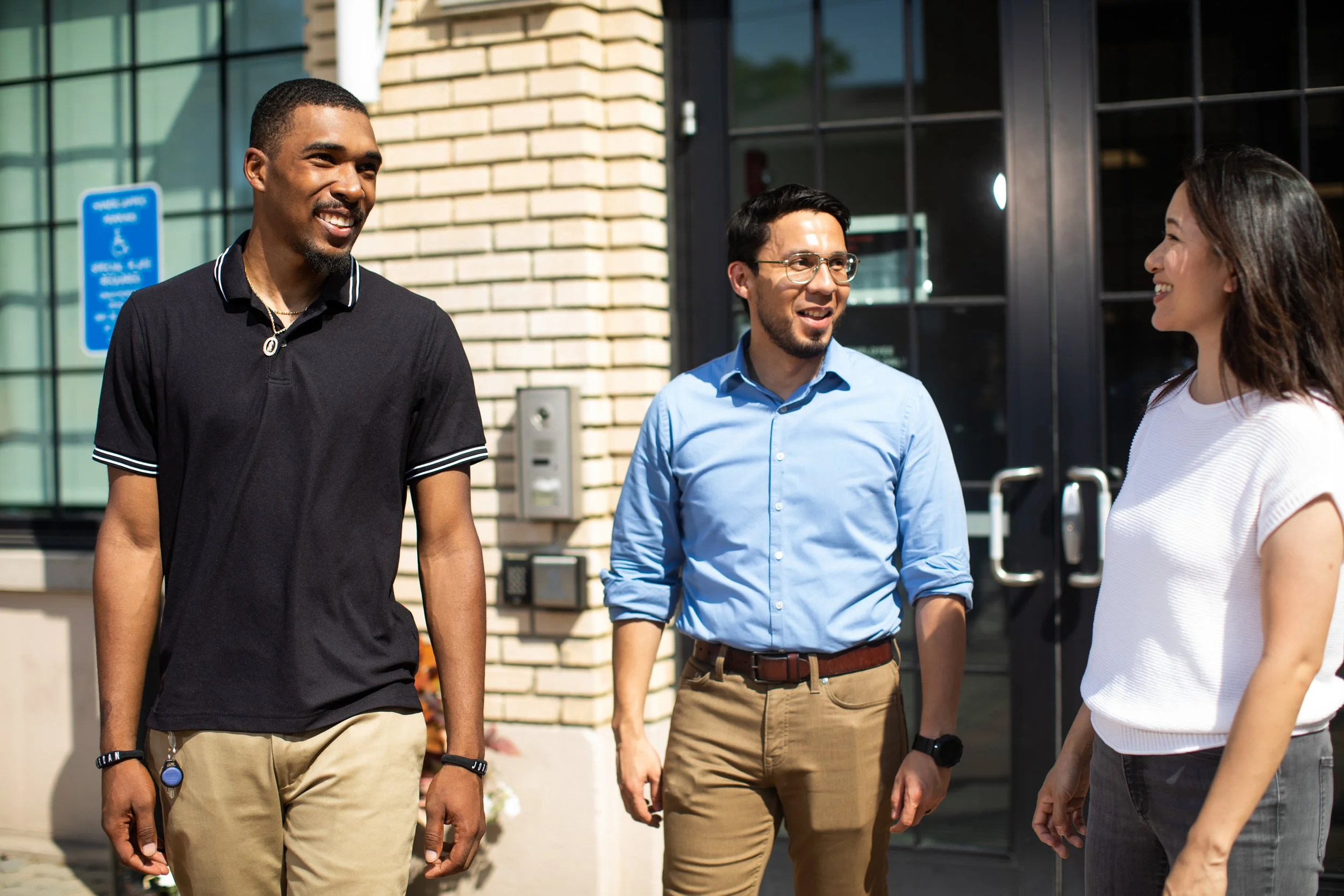 Three people, two men and one woman, are standing outdoors and engaging in conversation, smiling, in front of a building with a brick exterior and large glass windows.
