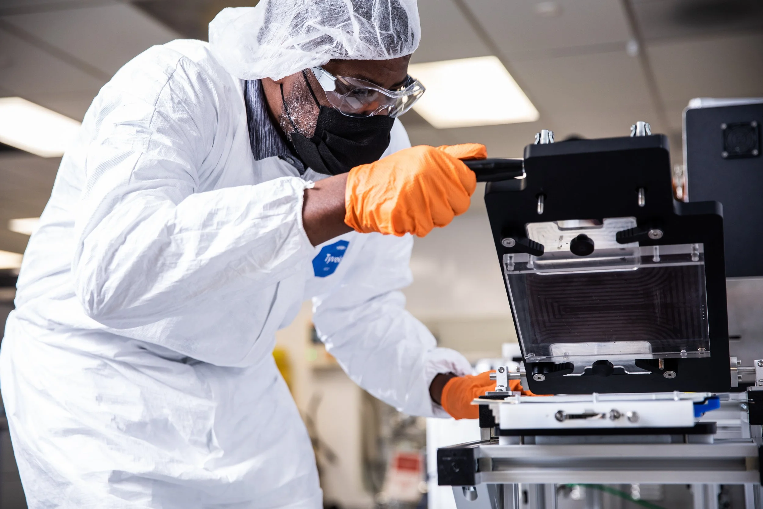 A scientist wearing protective gear, including a hair cover, glasses, a face mask, and orange gloves, is working with laboratory equipment.