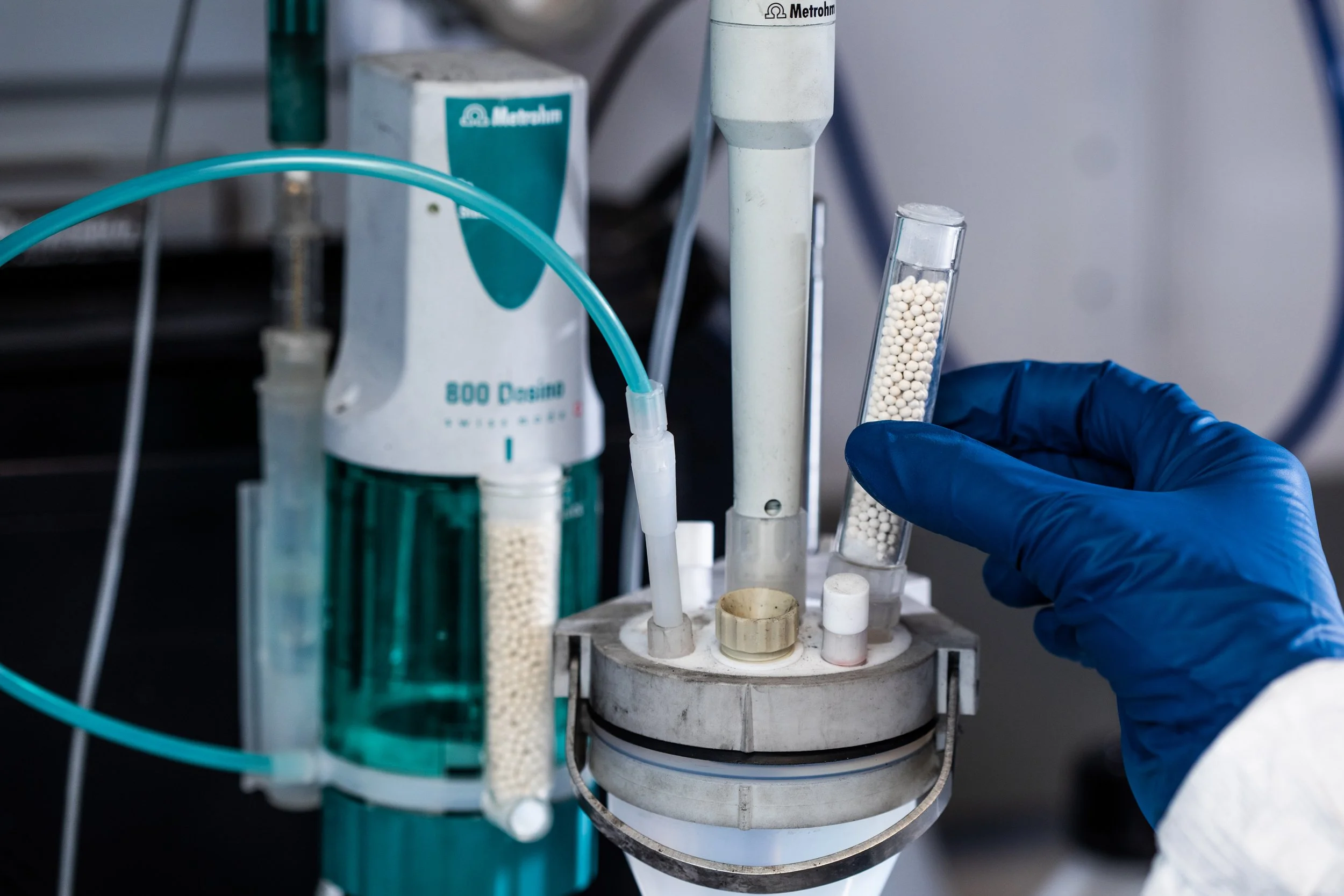 Close-up of laboratory equipment with a gloved hand holding a test tube filled with small white pellets, surrounded by various tubes, and machinery used for scientific experiments.