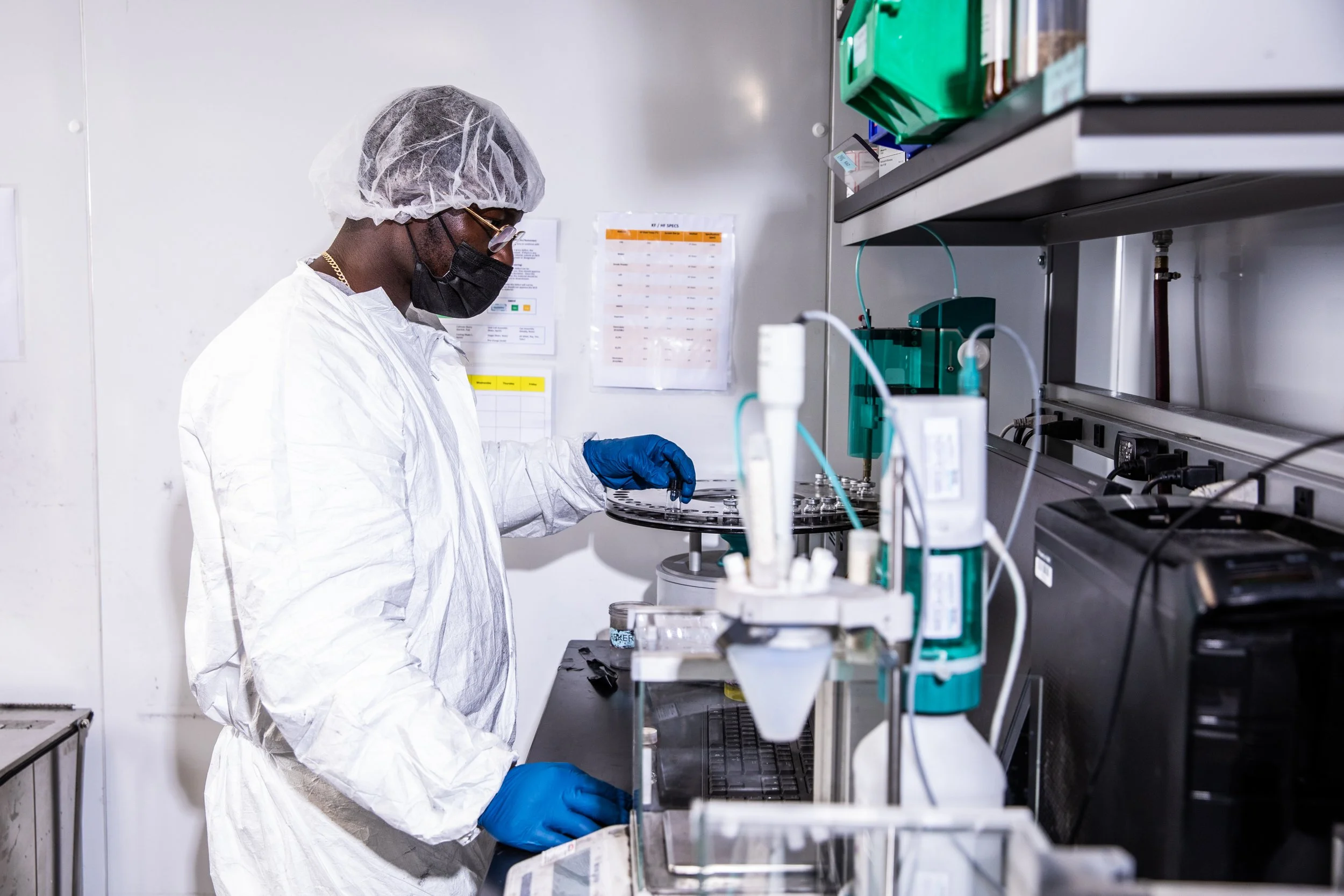A scientist wearing a white lab coat, hairnet, mask, glasses, and blue gloves working in a laboratory with scientific equipment and supplies.