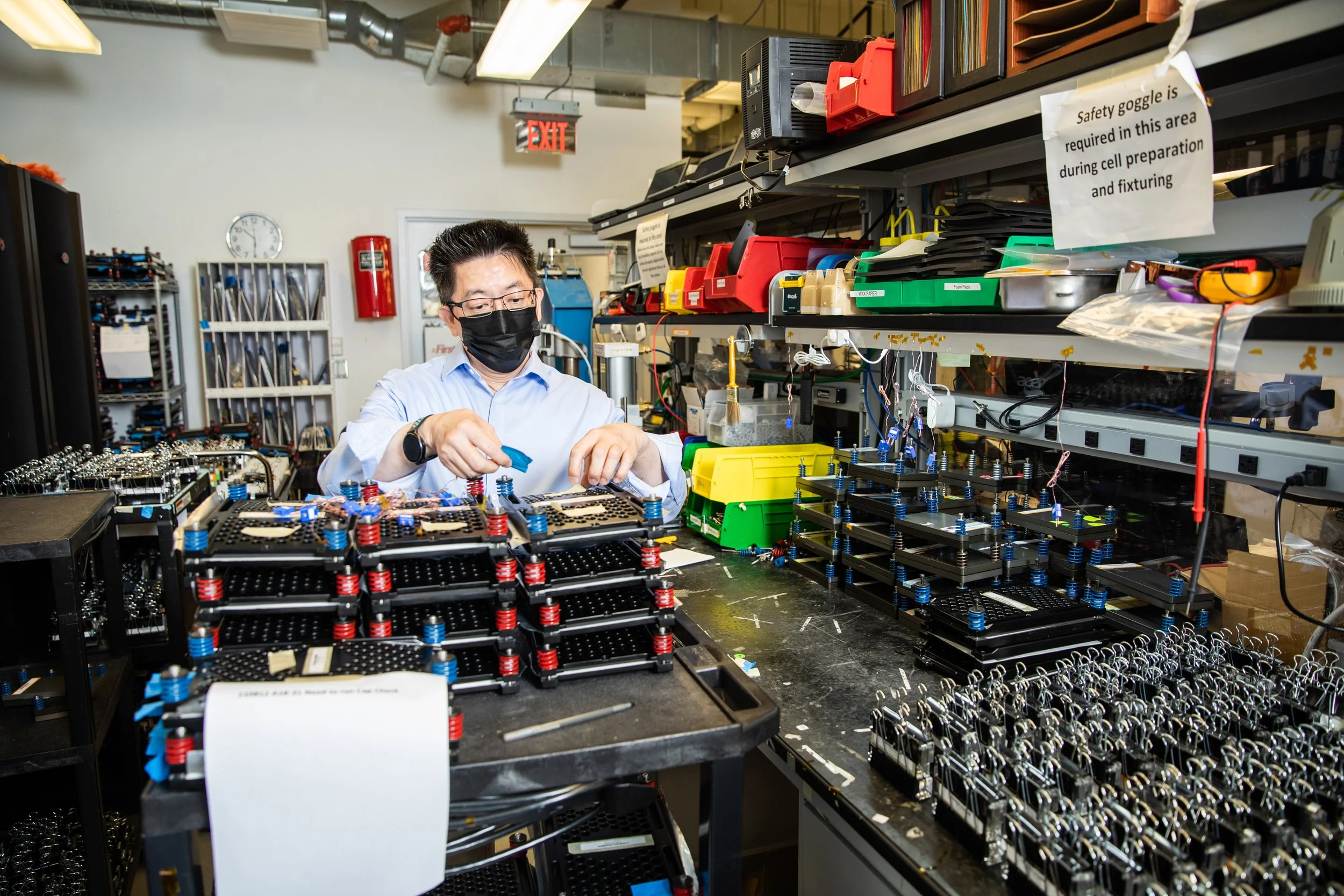 A person wearing glasses and a black face mask working on assembly of electronic components with organized tools and shelves around in a workshop.