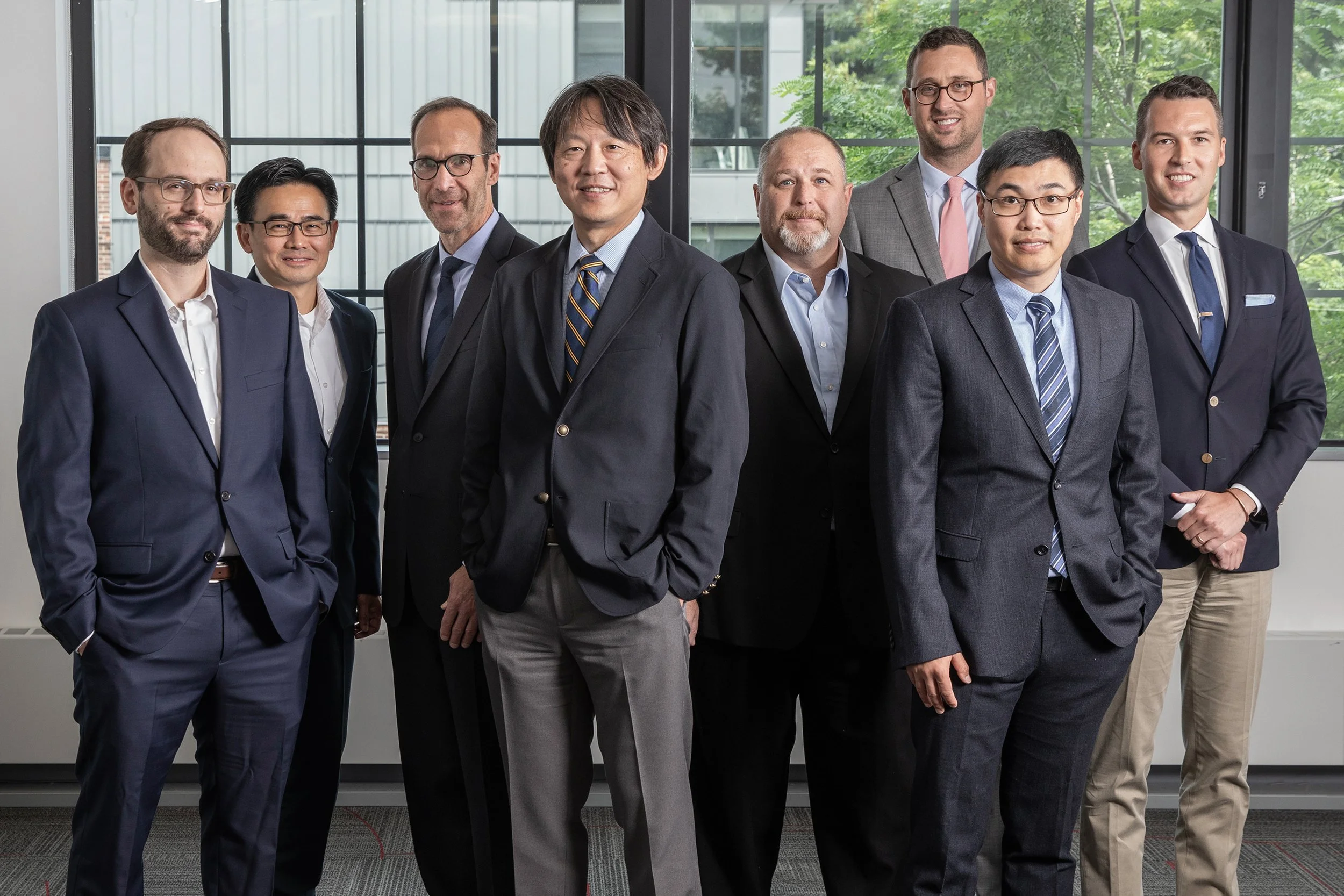 A diverse group of nine well-dressed men posing for a photo in a modern office with large windows and green trees outside.