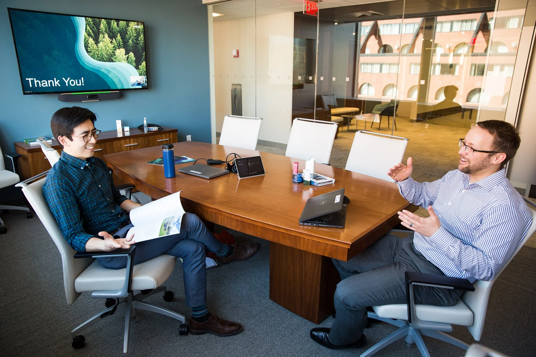Two men having a conversation in a conference room with a large wooden table, white chairs, laptops, and an extra monitor. One man is wearing a striped shirt and glasses, gesturing with his hands, while the other man, also with glasses, is smiling an