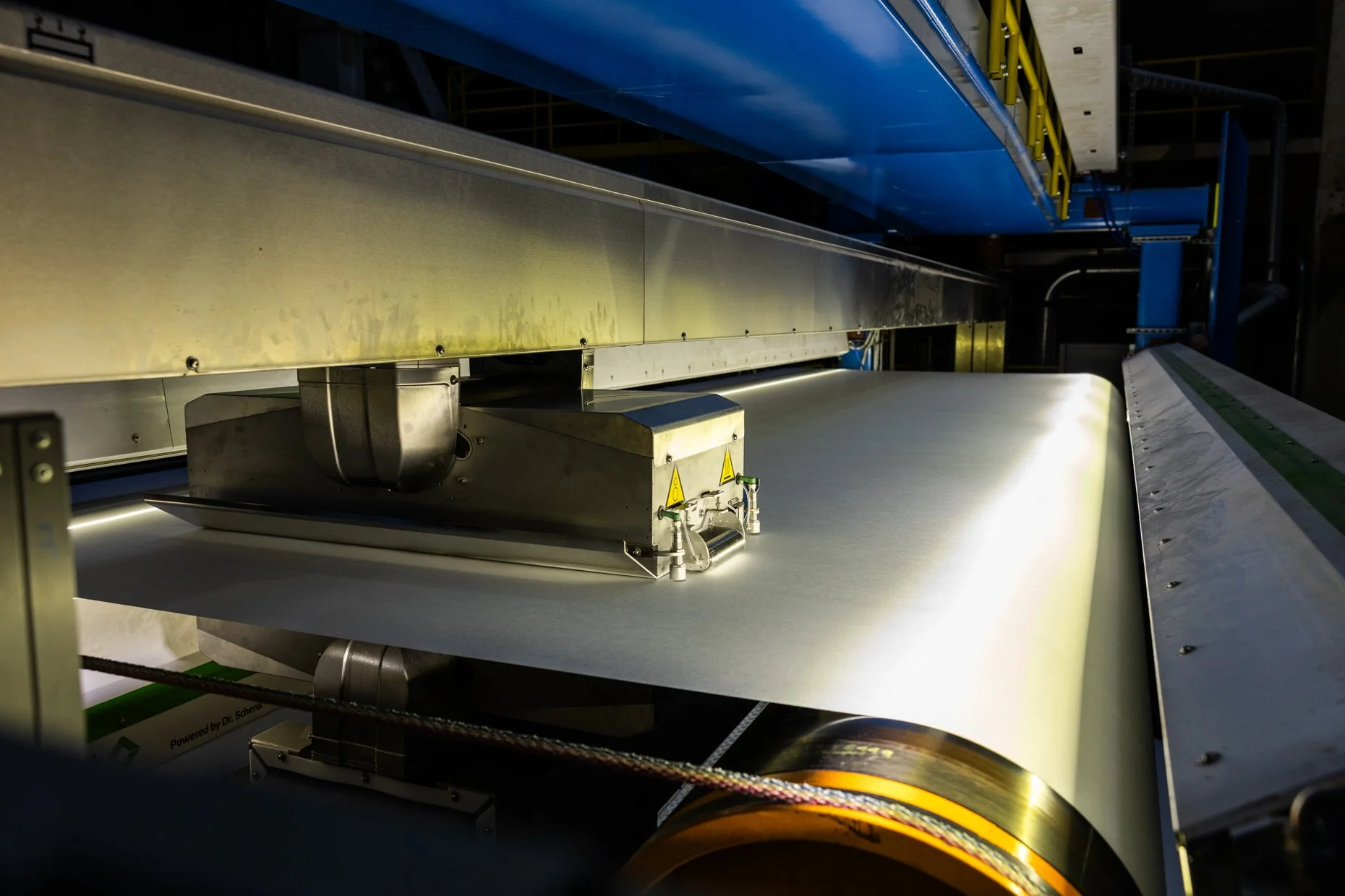 A large industrial machine with metallic and yellow components, including a conveyor belt, pipes, and safety warnings.