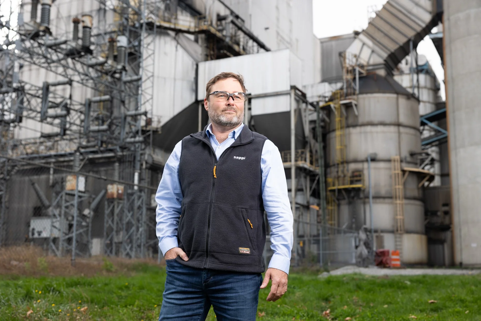 A man standing outdoors in front of industrial equipment and structures, wearing glasses, a light blue shirt, and a black vest.