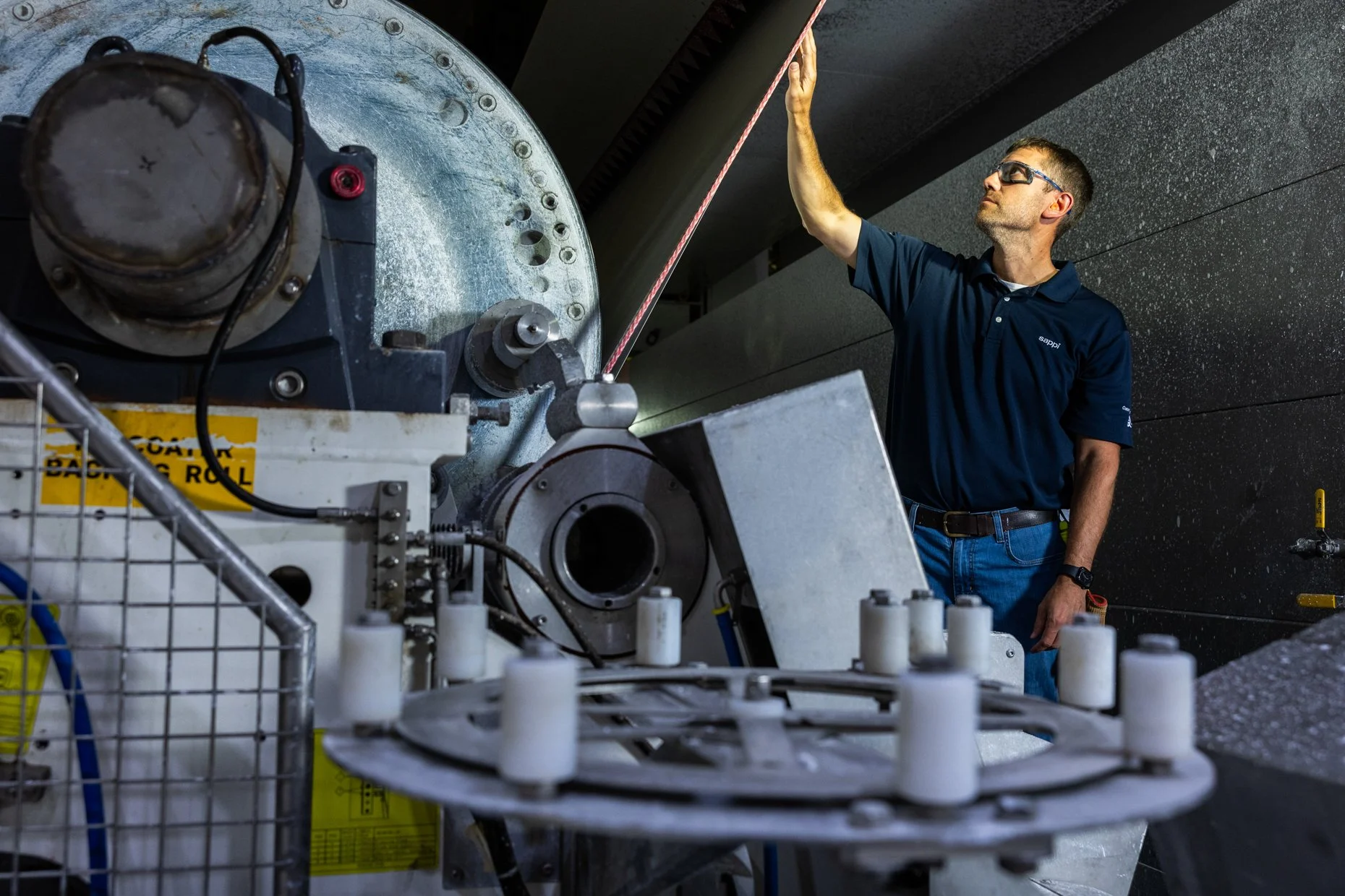 A man wearing safety glasses and a navy blue polo shirt is working with industrial machinery, which includes a large round metal component with multiple bolt holes. The setting appears to be an industrial or manufacturing environment.