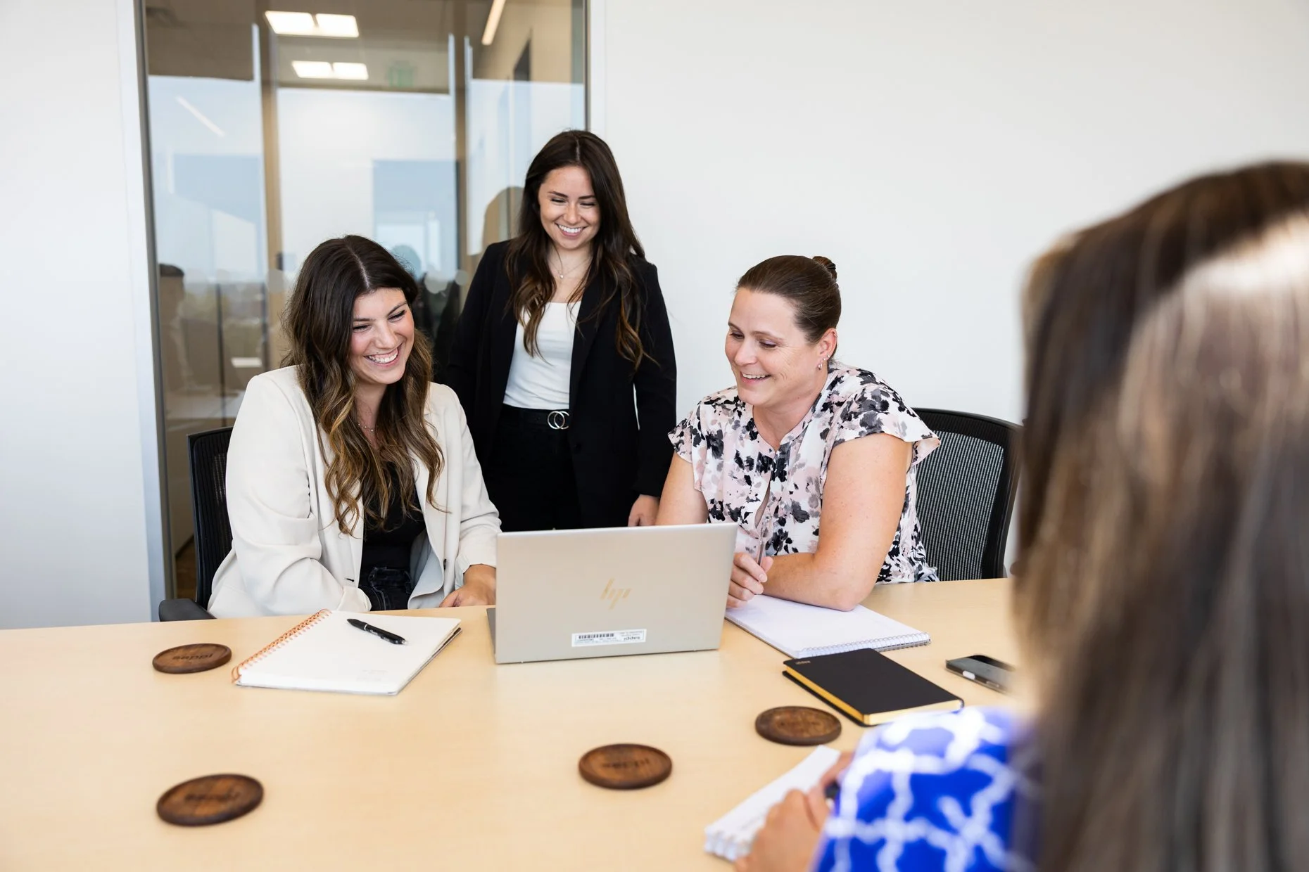 Four women in a meeting room look at a laptop screen and smile, while another woman stands and looks on. The table has notebooks, a smartphone, and coasters.