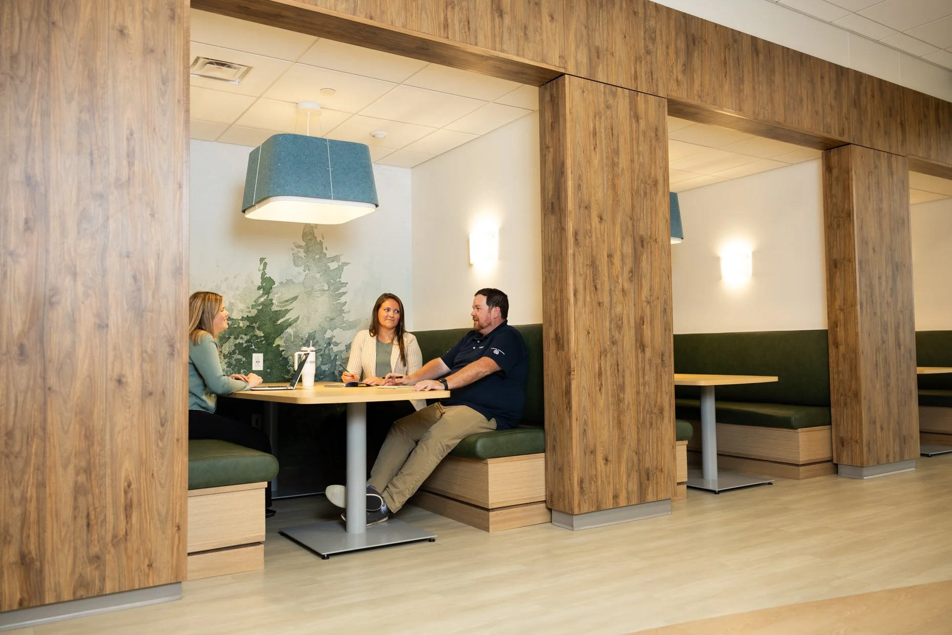 Three people sitting in a booth at a restaurant or cafe, engaged in conversation. The booth is surrounded by wooden partitions, with pendant lights hanging from the ceiling. The walls have a mural of a pine tree, and there is a laptop and a water bot