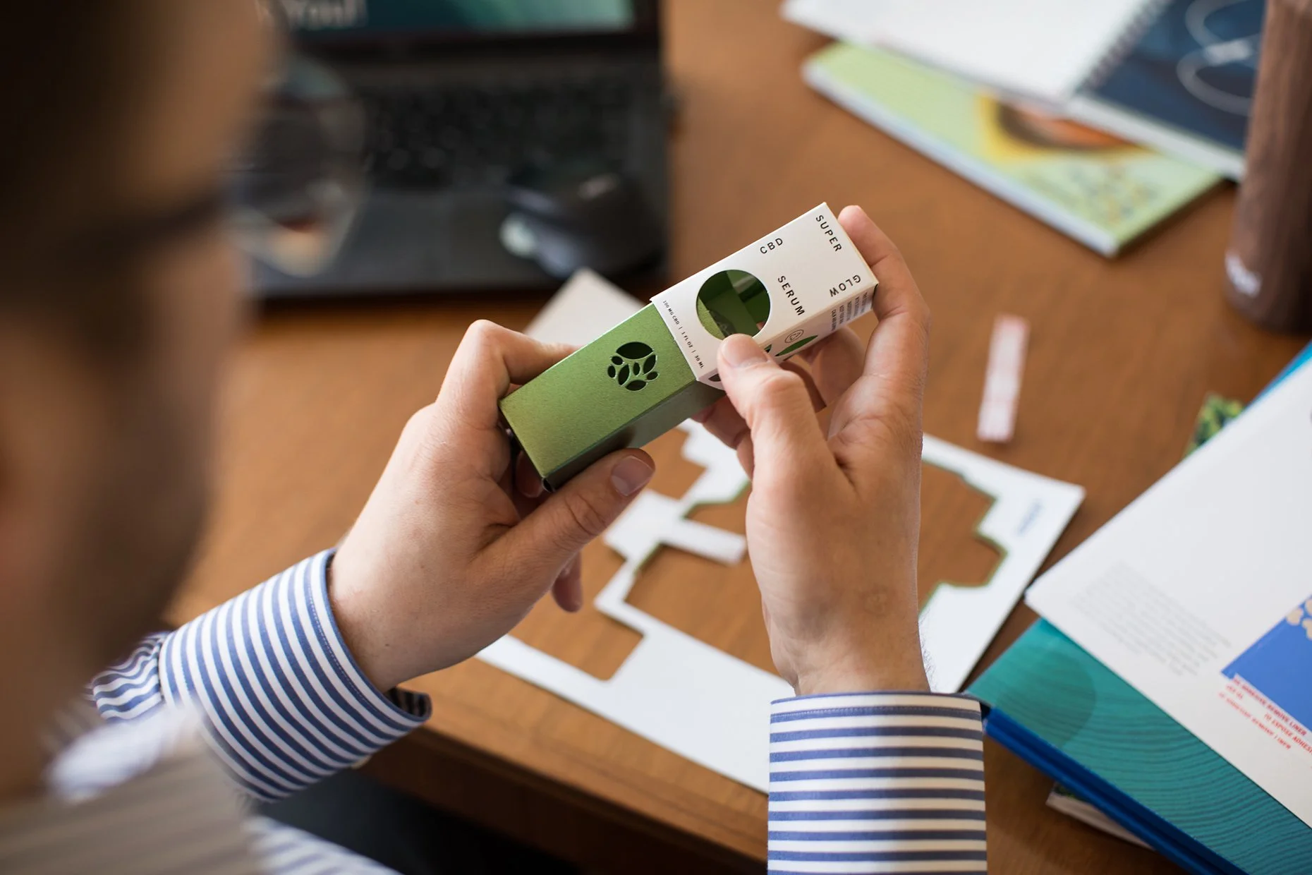 A person holding a rectangular box labeled 'CBD Super Serum' with a green and white design, sitting at a wooden desk with various papers, notebooks, and a laptop.