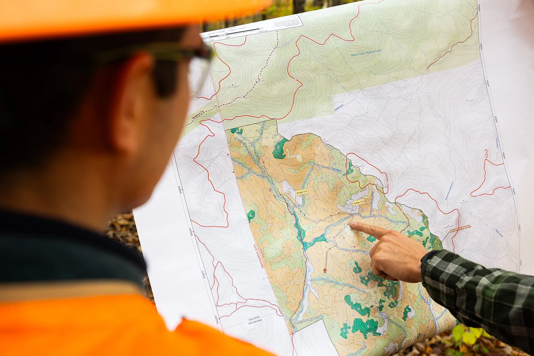 A person in an orange safety vest and a person in a plaid shirt pointing at a detailed topographic map, likely discussing land features and boundaries.