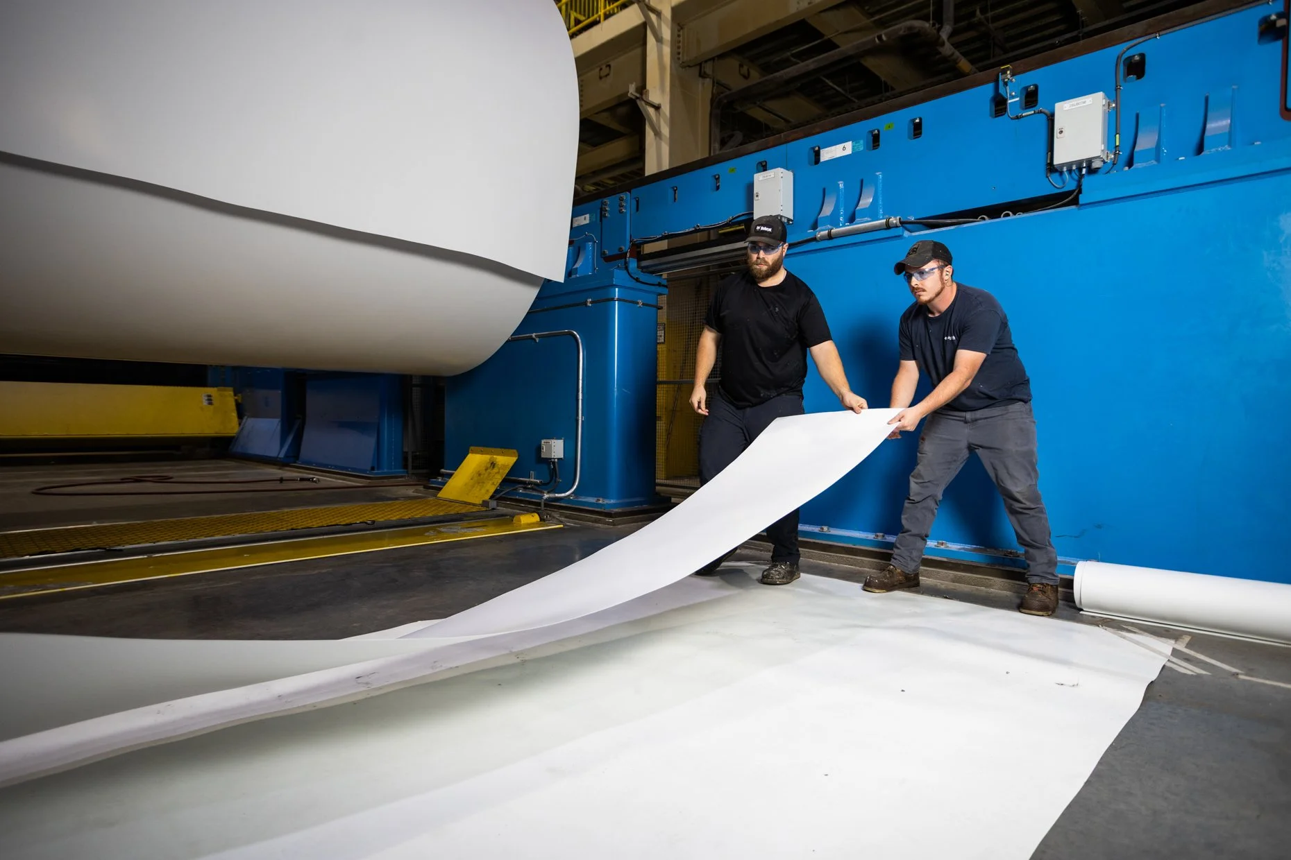 Two workers in safety glasses and hats operate a large industrial printing machine, unrolling a long sheet of white material in a manufacturing facility.