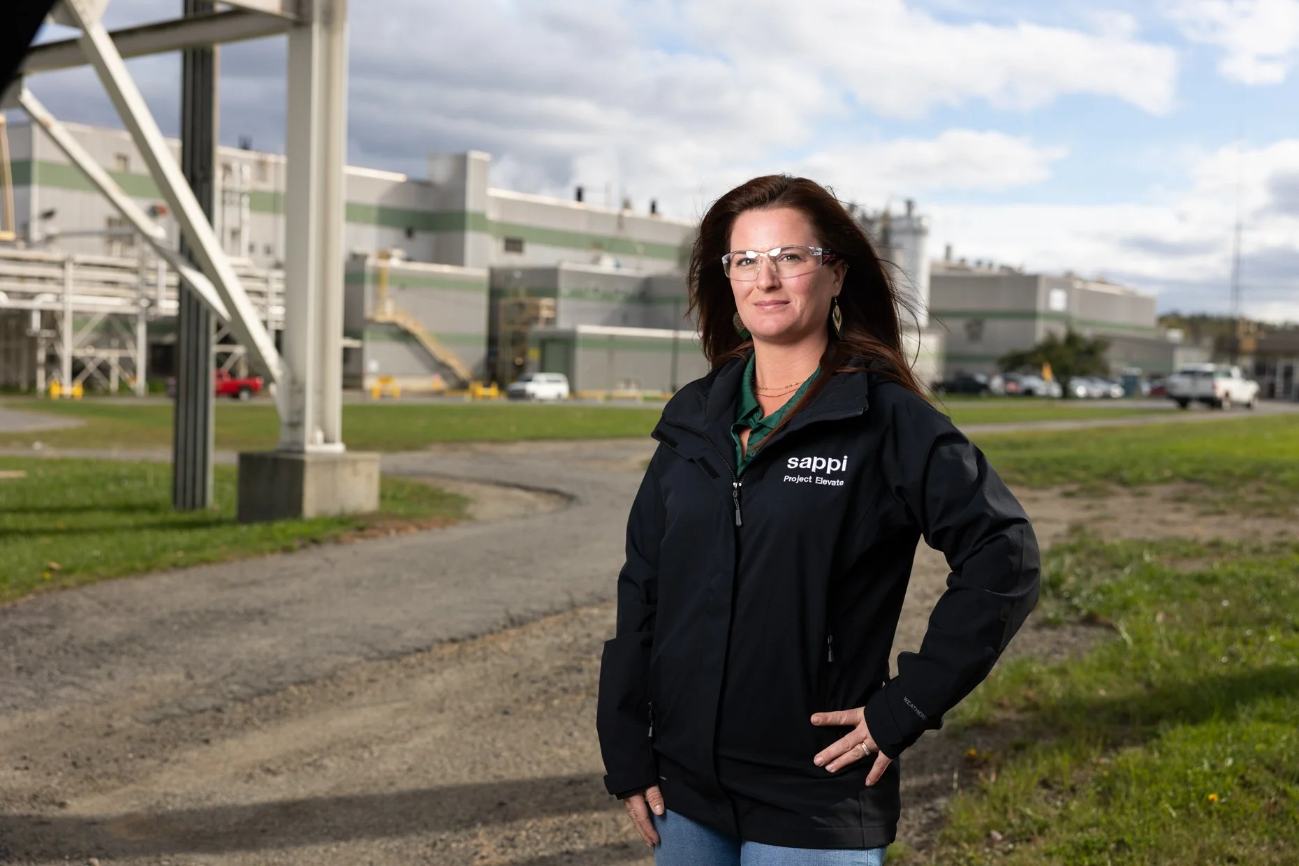 A woman with long brown hair, glasses, and earrings standing outdoors in front of a building with a cloudy sky in the background. She is wearing a black jacket with the logo 'sappi' and the words 'Project Elevate' on it.