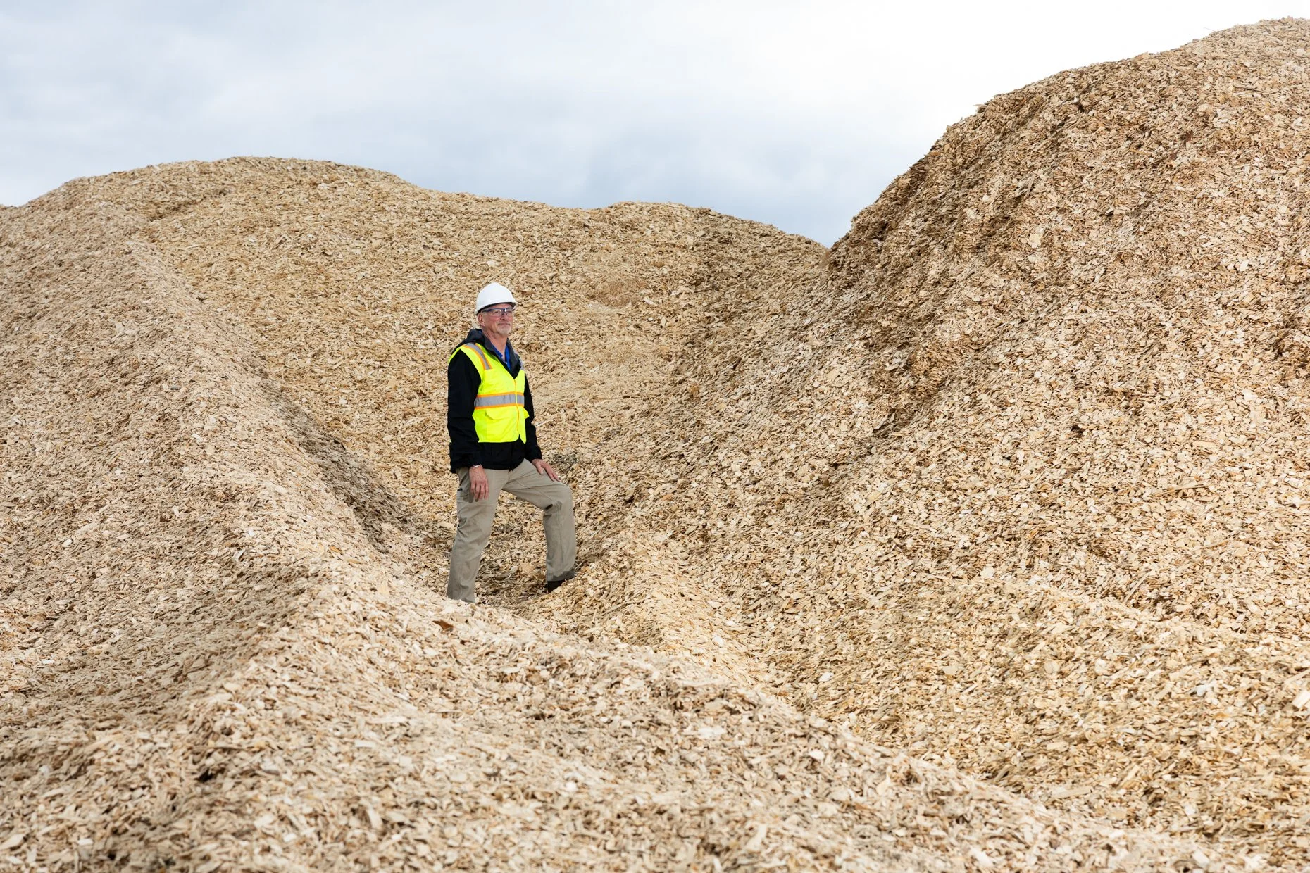 A man wearing a yellow safety vest, khaki pants, and a white hard hat standing on a large pile of wood chips in an outdoor setting.