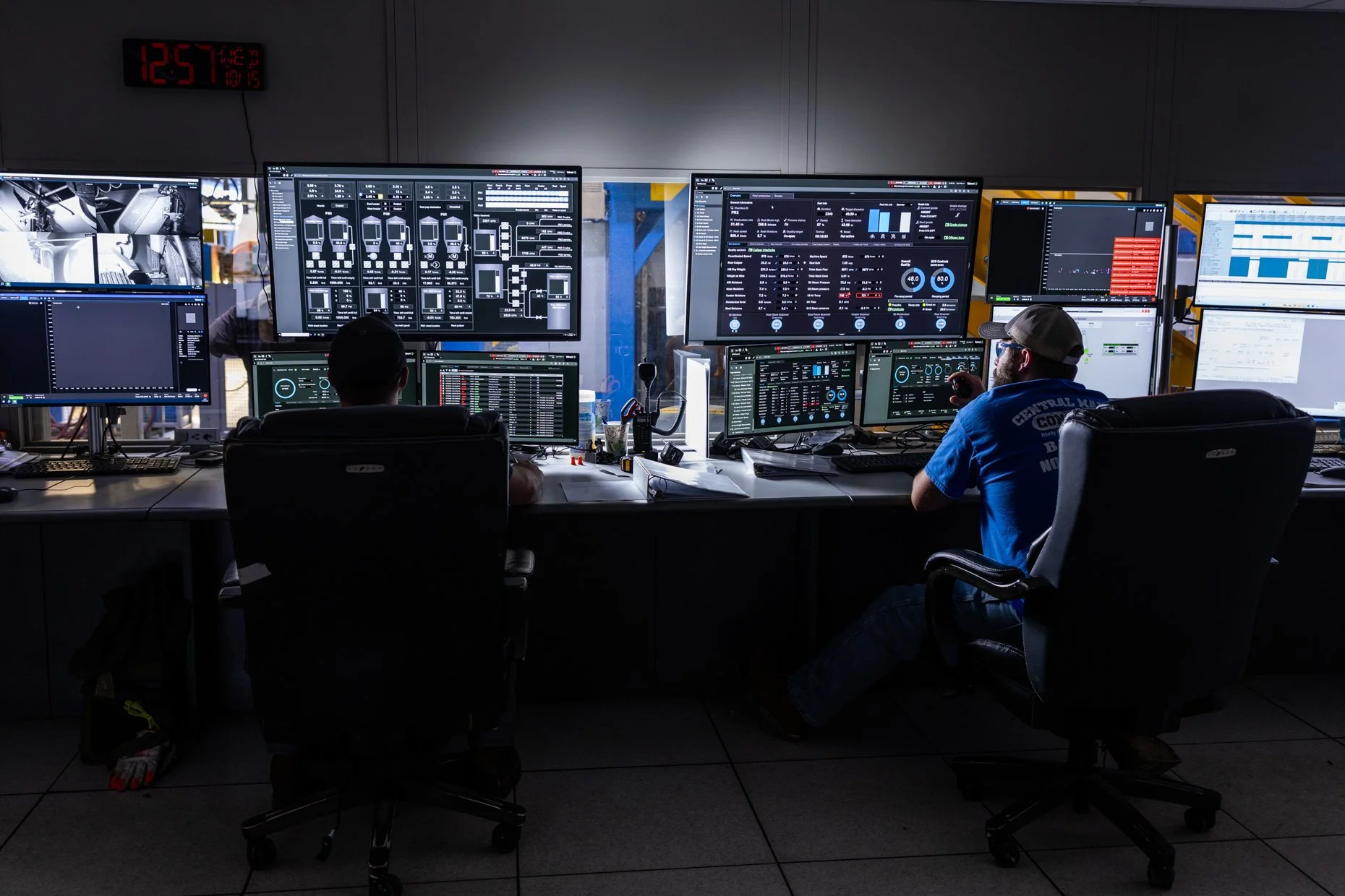 A control room with multiple monitors displaying technical data and diagrams. Two people are seated at the desk working, one wearing a baseball cap and glasses. A digital clock on the wall shows the time as 12:57.