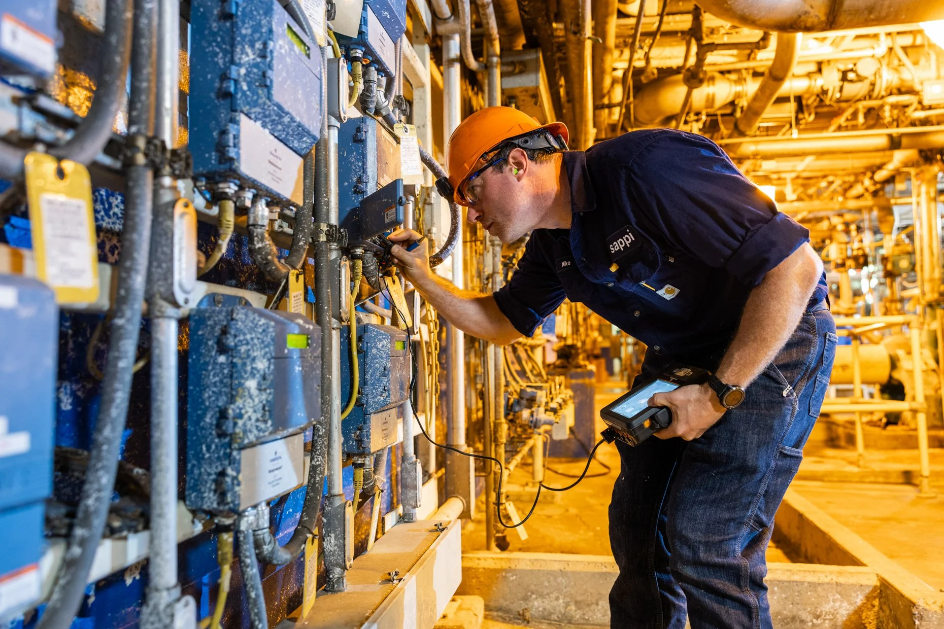 A worker wearing a hard hat and safety glasses inspects electrical equipment with a diagnostic device in an industrial setting with pipes and machinery.