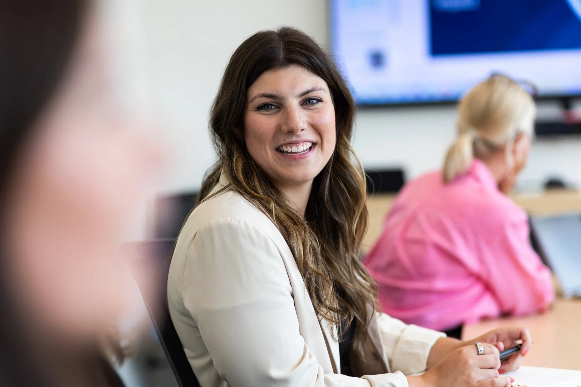 A woman with long brown hair smiling and holding a smartphone, sitting in a conference room with two other women, one wearing a pink shirt and glasses, and a large monitor displaying a blue screen in the background.