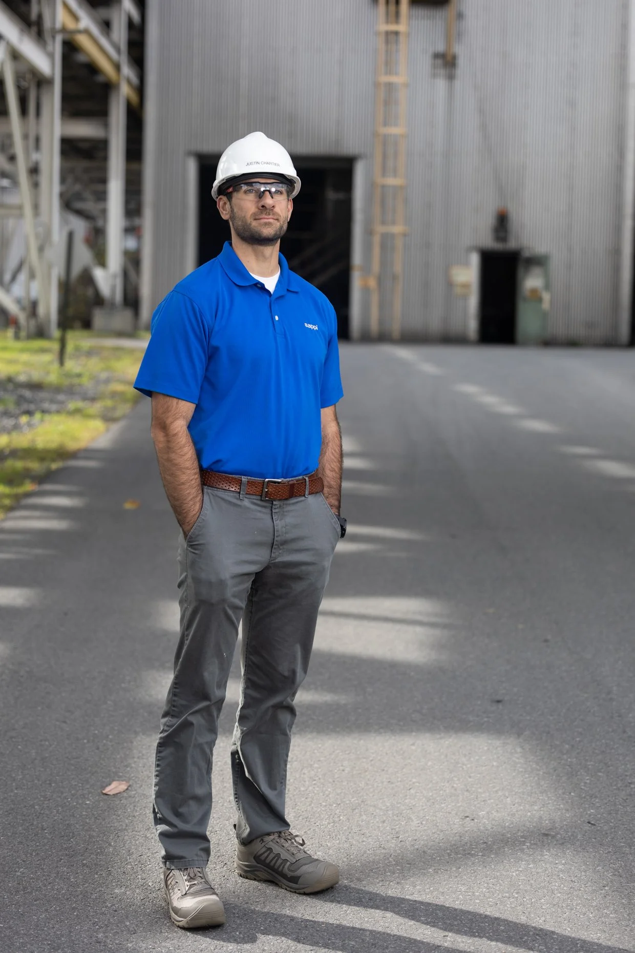 A man wearing safety glasses, a white safety helmet, a blue polo shirt, gray pants, and gray athletic shoes standing outside in an industrial area with a building and construction equipment in the background.