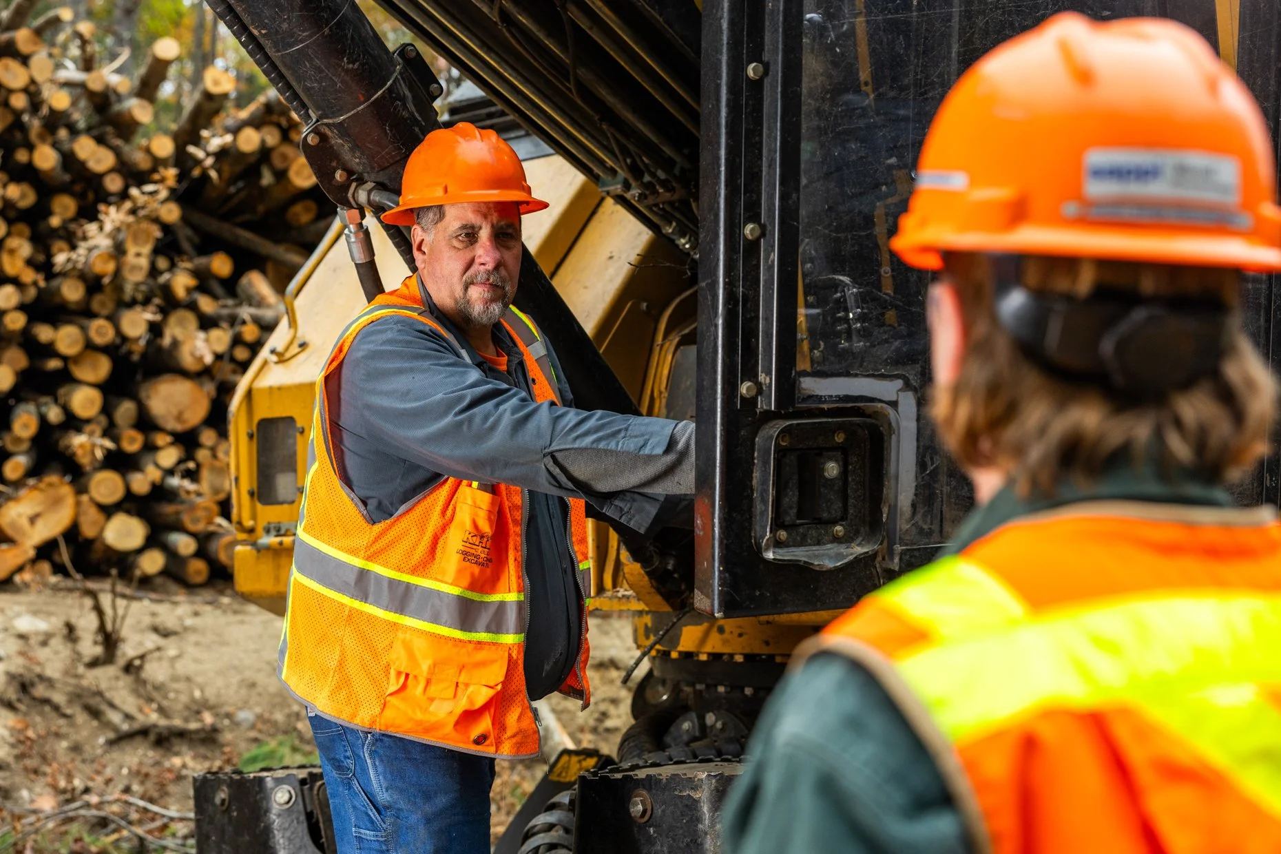 Two construction workers wearing orange safety helmets and reflective vests working on a yellow logging vehicle. One worker with a beard looks towards the camera while adjusting or inspecting the machine, with a large pile of cut logs in the backgrou