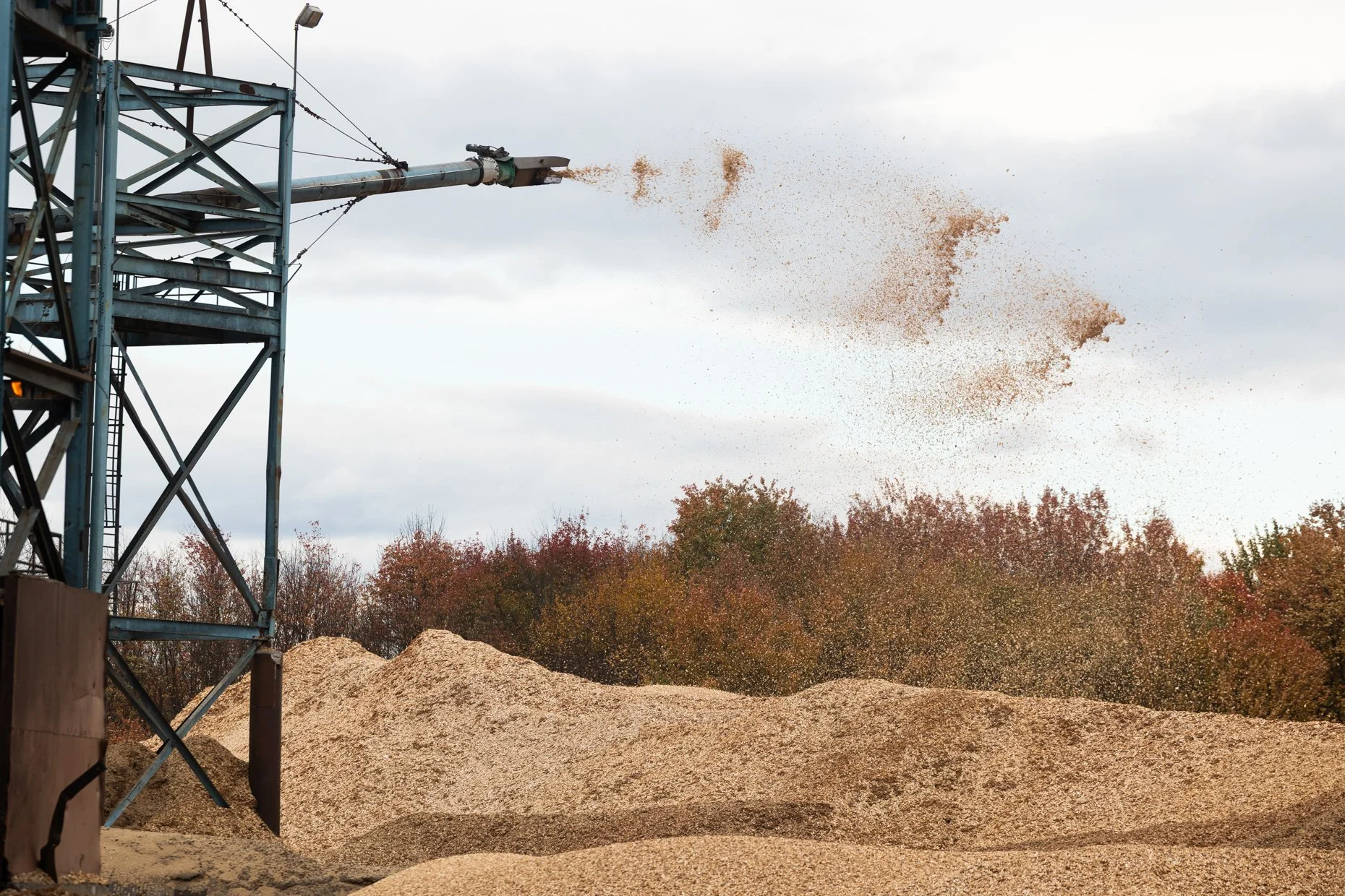 A large industrial dust or grain loading machine releasing particles into the air, with a steel tower structure on the left and piles of granular material on the ground, in a rural outdoor setting with trees in the background.