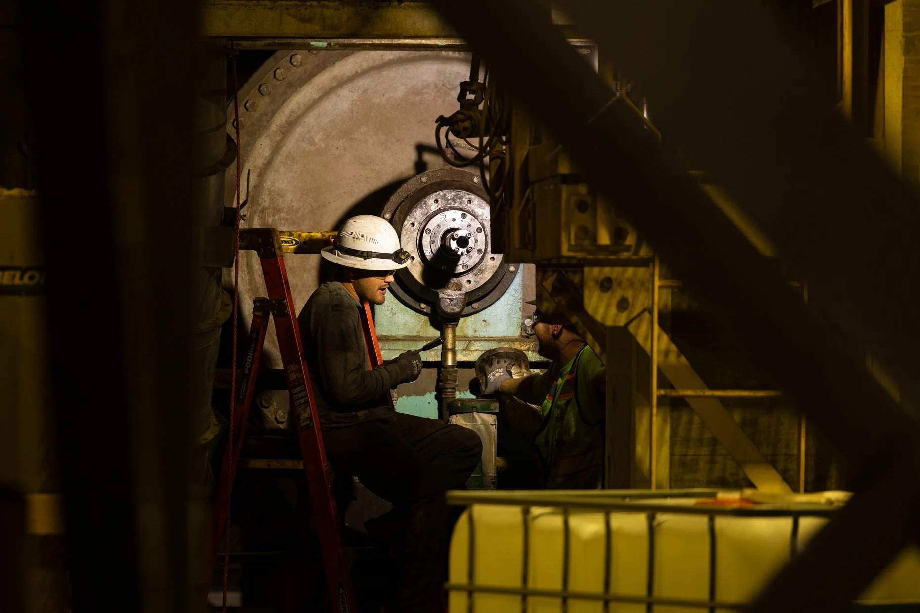 Two workers wearing safety helmets and gloves are working on a large, industrial machine in an underground tunnel or mine. One worker is seated and the other is standing, both focused on the task.