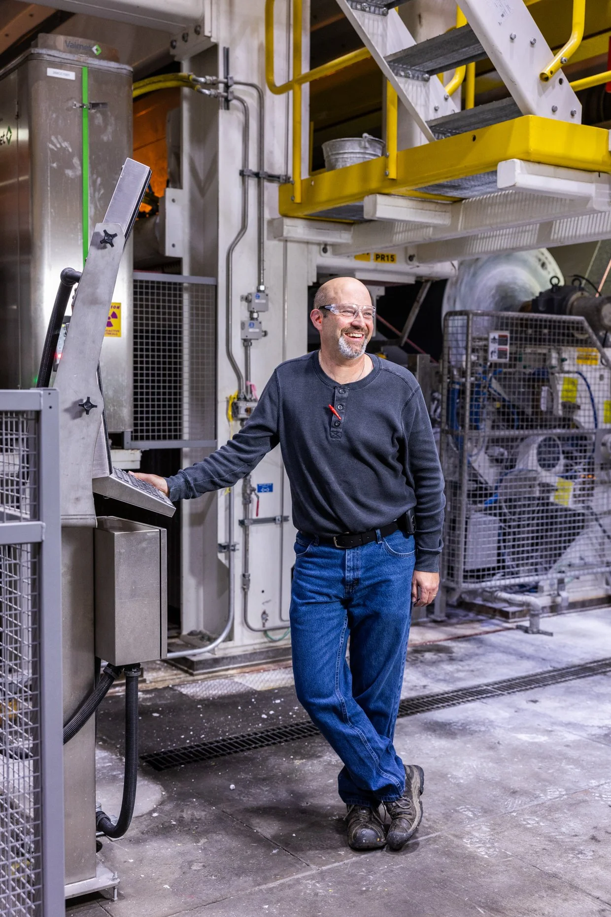 A man in safety glasses and casual clothing standing in an industrial setting, smiling while holding a control panel. The background includes industrial equipment, piping, and a yellow staircase.