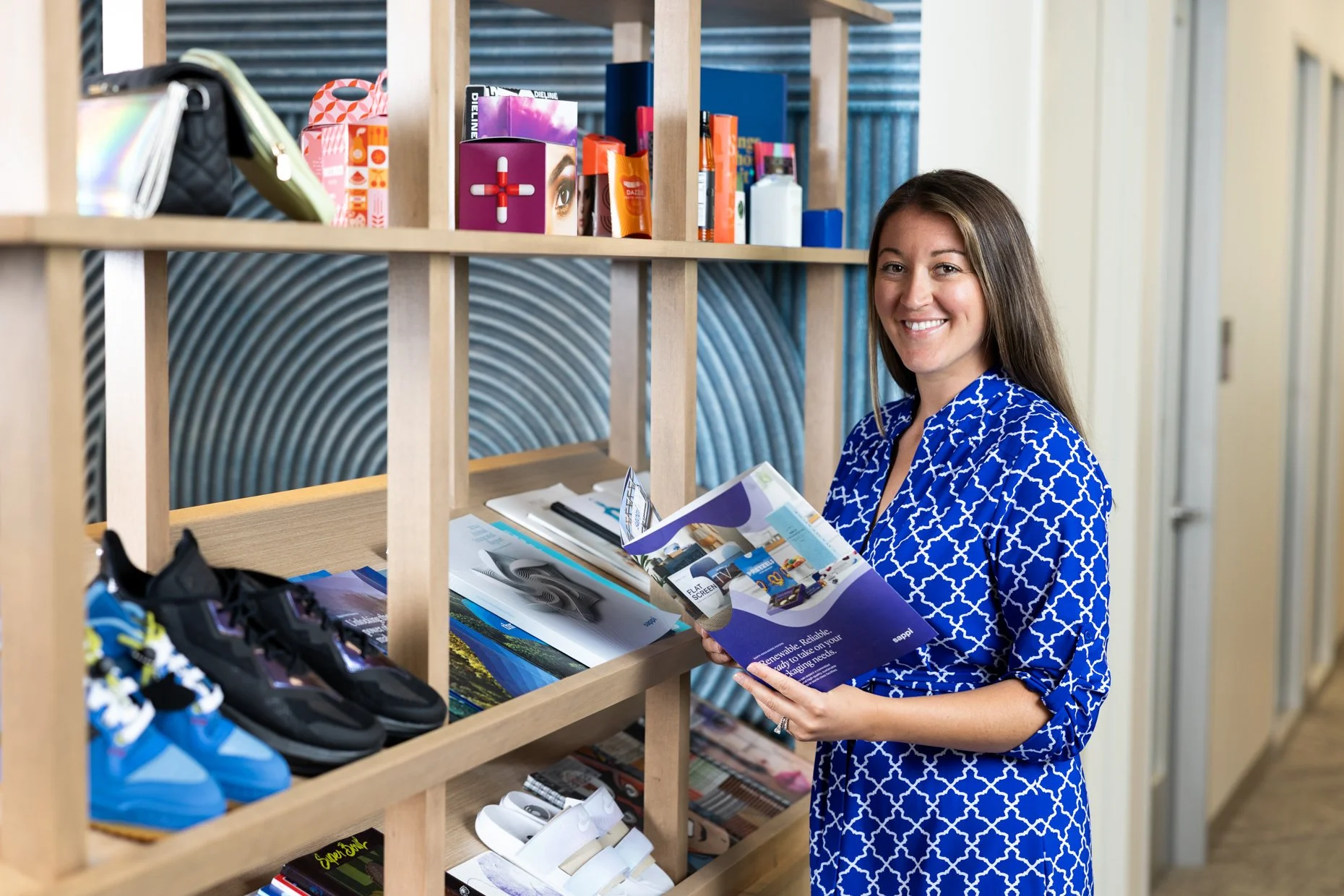 A woman in a blue patterned shirt holding a brochure, smiling, standing behind a wooden shelf with sports shoes, magazines, and other products. A background wall has a textured design.