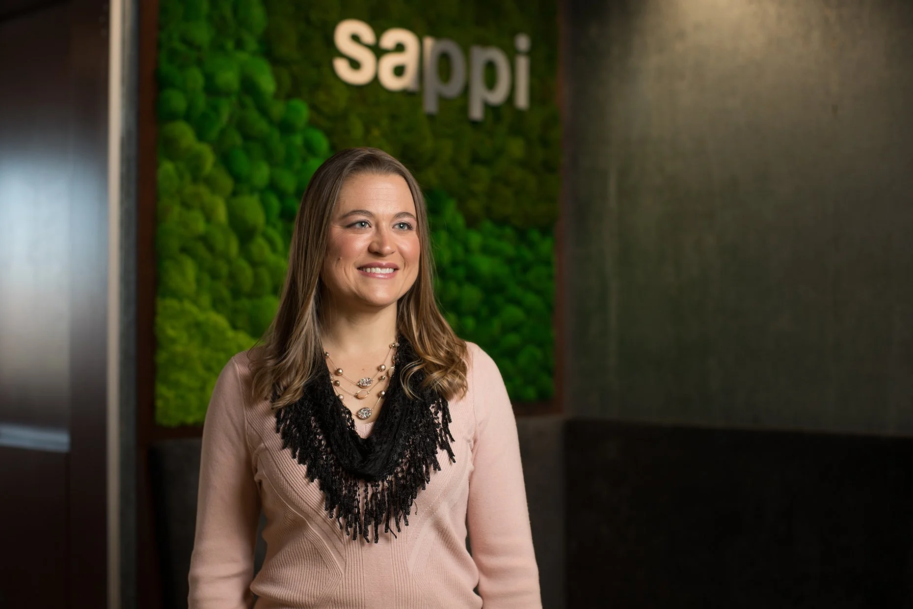 A woman with long, wavy light brown hair, smiling, standing in front of a green moss wall with the word 'Sappi' on it. She is wearing a pink sweater with black lace detail and layered pearl necklaces.