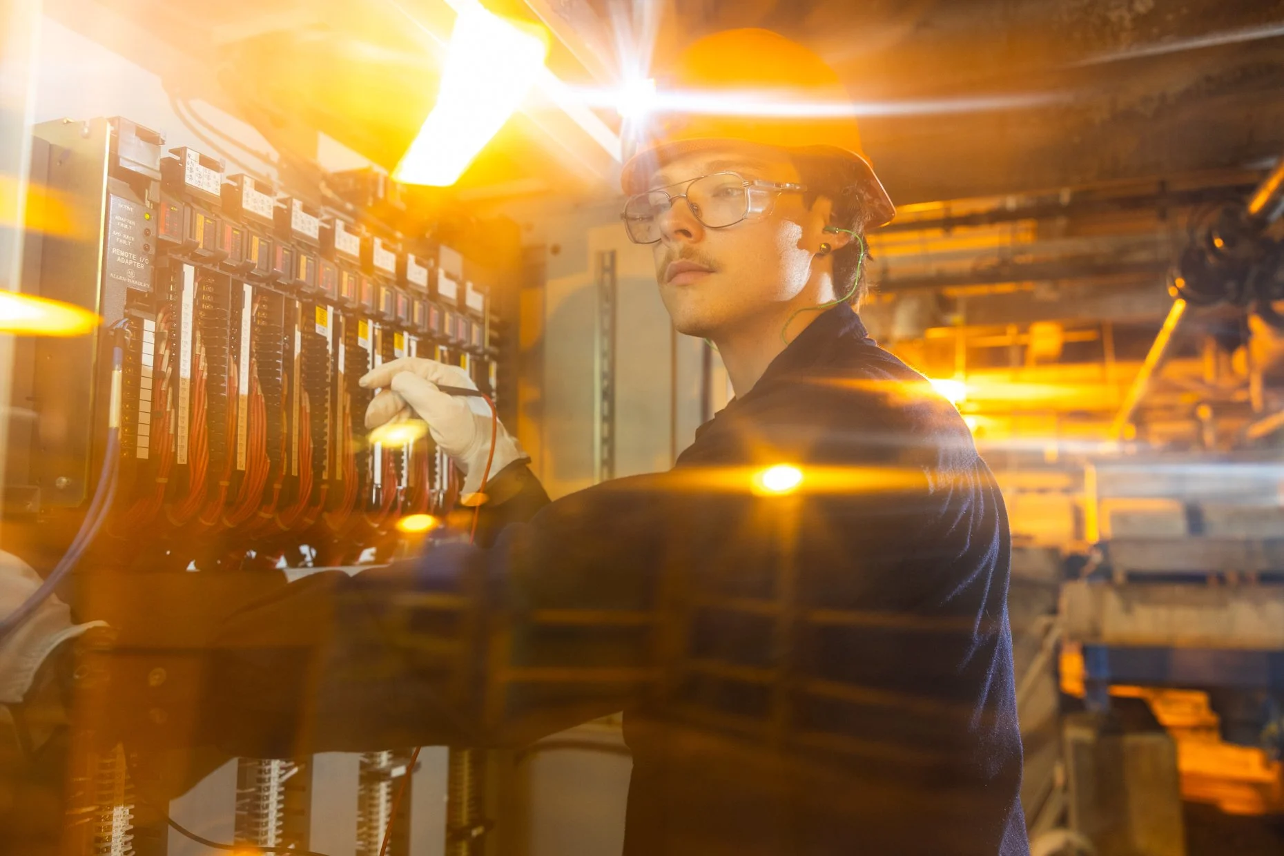 Technician working on electrical equipment in a server room, wearing safety helmet, glasses, and gloves, illuminated by yellow light.